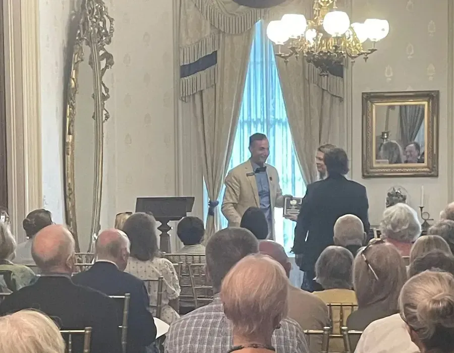 Audience seated in a formal room while two men speak near a piano and tall window