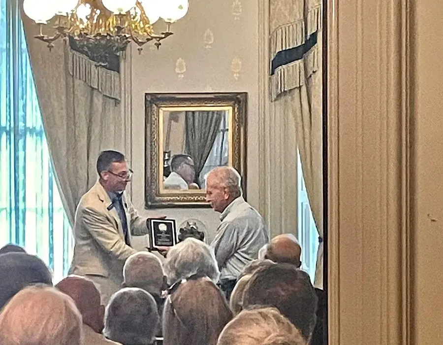 Two men in suits shake hands during a formal presentation in a decorated room, seen over an audience.