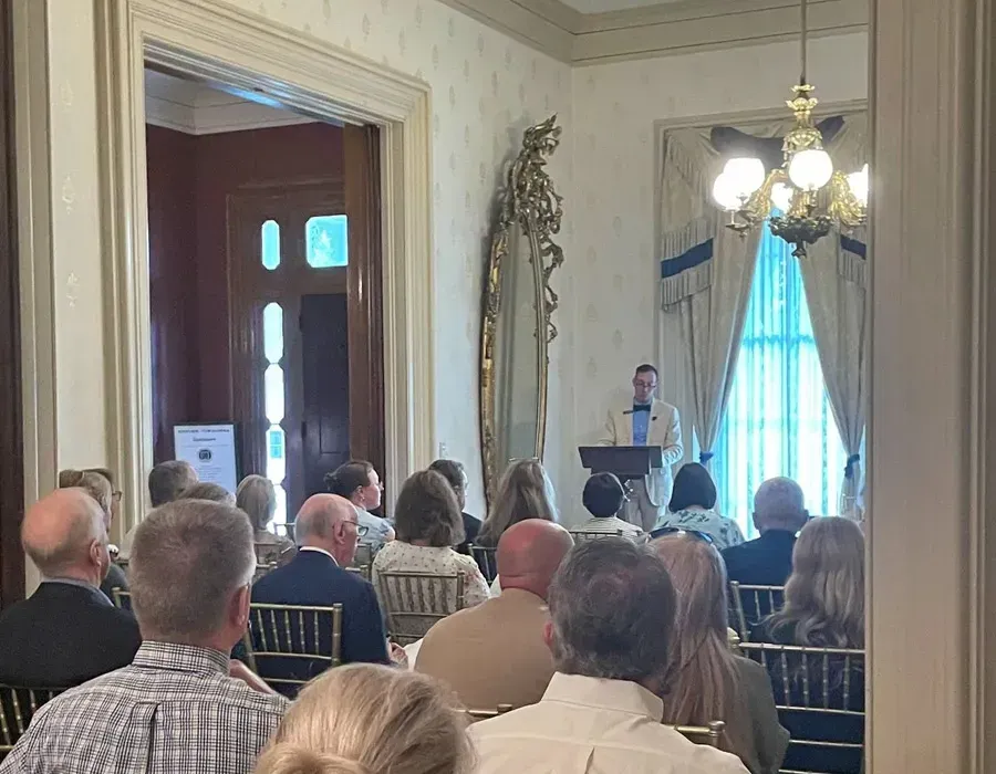 Audience seated in a formal room listening to a speaker at a podium near a bright window.
