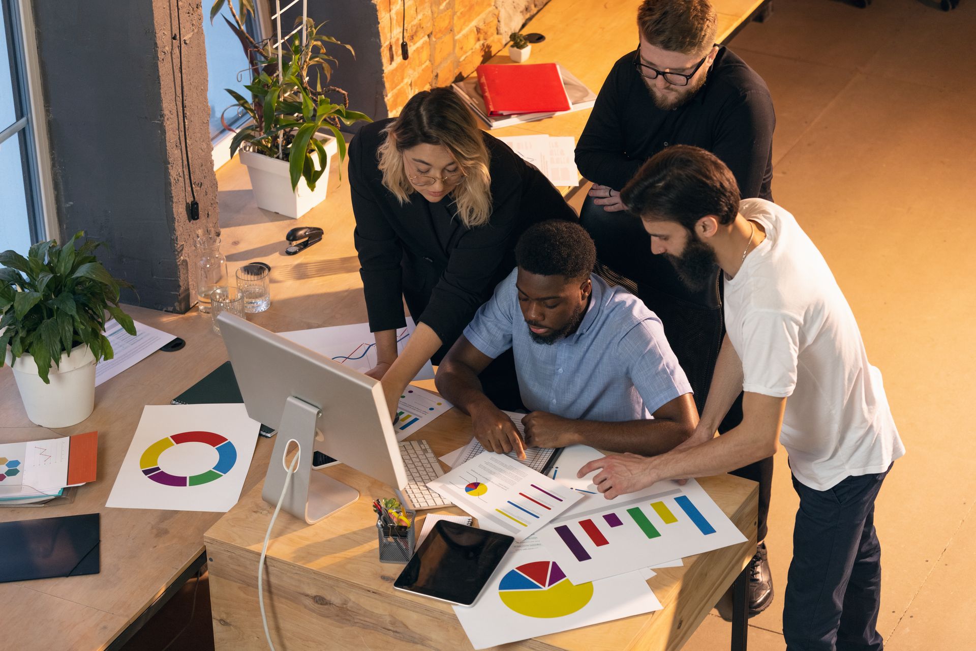 Four coworkers collaborate around a desk, reviewing colorful data charts and graphs on a computer monitor and paper.