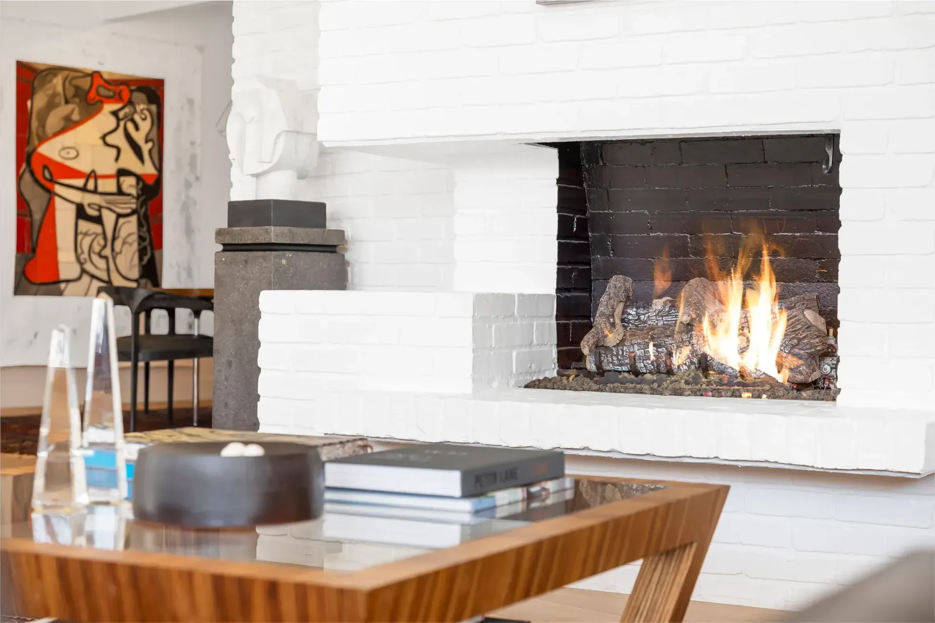 Modern living room with a lit fireplace, white brick wall, wooden coffee table, and abstract painting