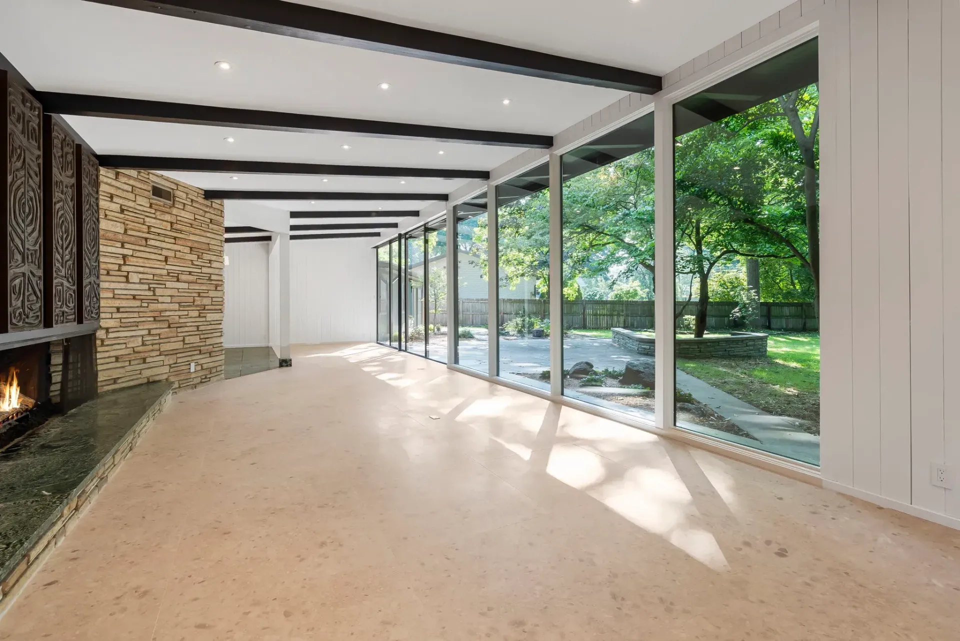 Modern open living room with fireplace, exposed beams, and floor-to-ceiling glass walls overlooking greenery