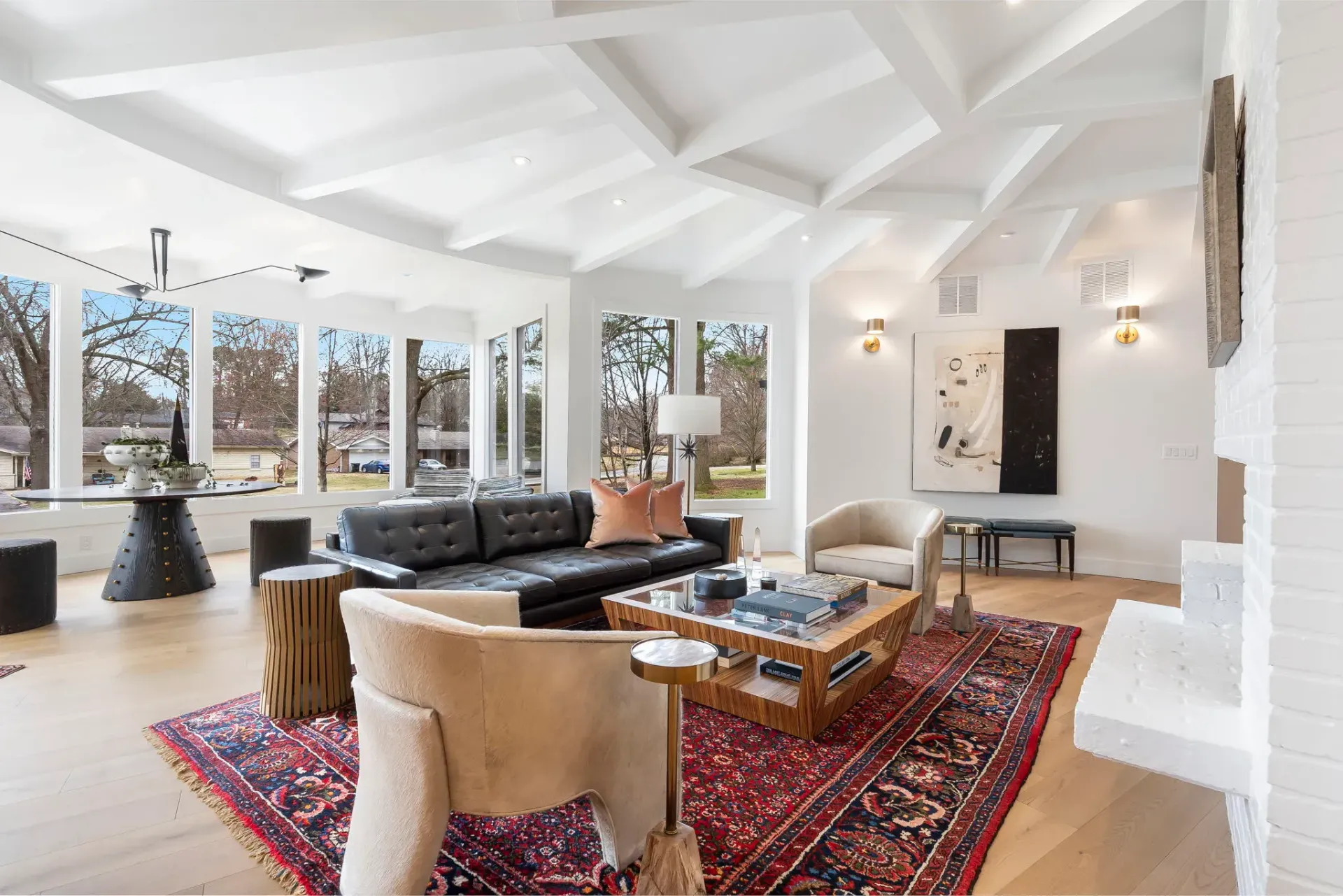 Bright living room with white beams, black sofa, tan chairs, red rug, and large windows.