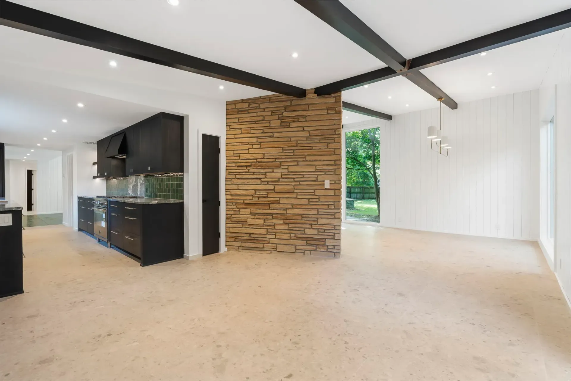 Spacious modern living room with exposed beams, stone fireplace, black kitchen cabinets, and beige carpet.