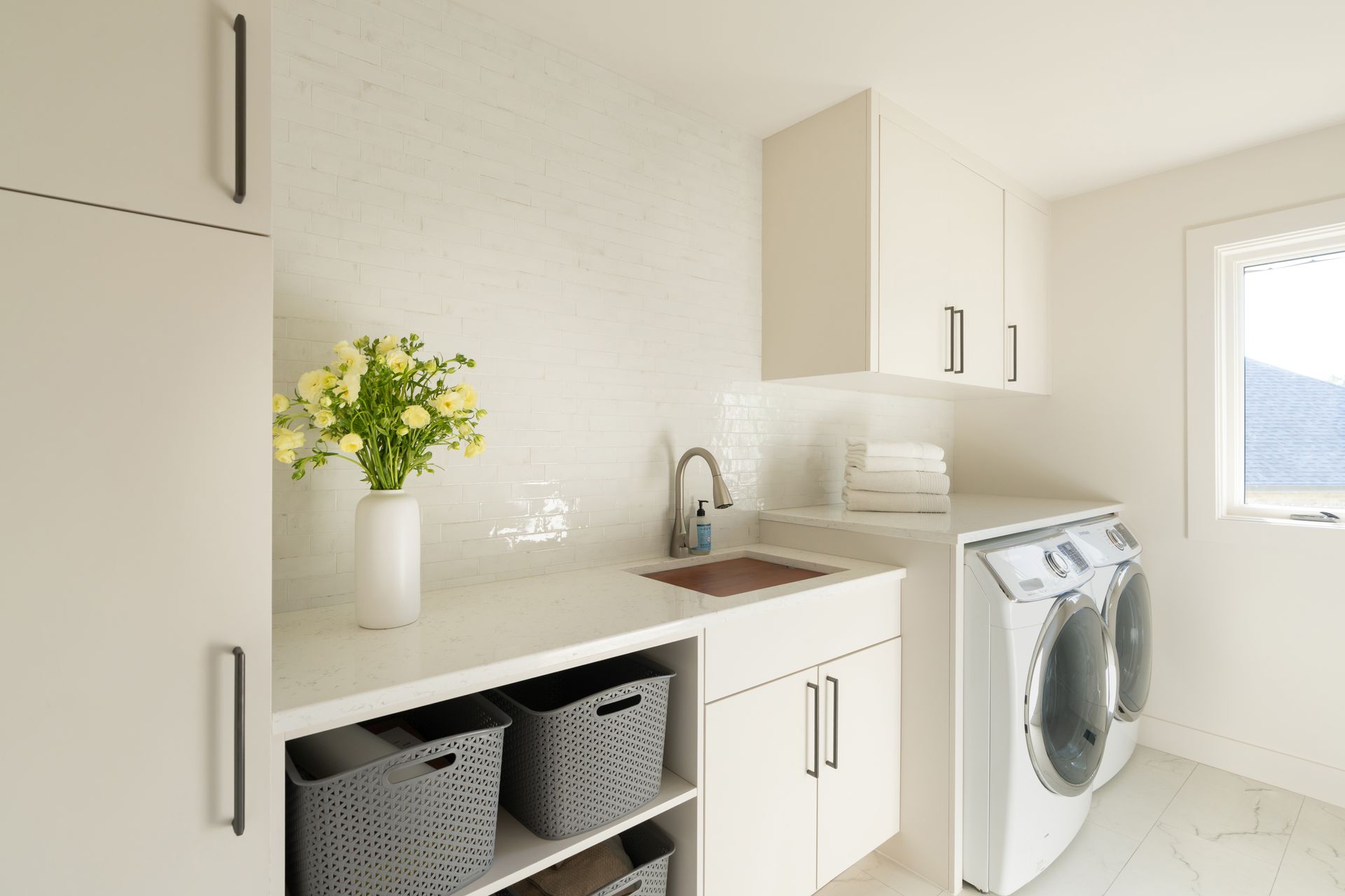 Bright laundry room with white cabinets, sink, washer, and storage baskets by a window.