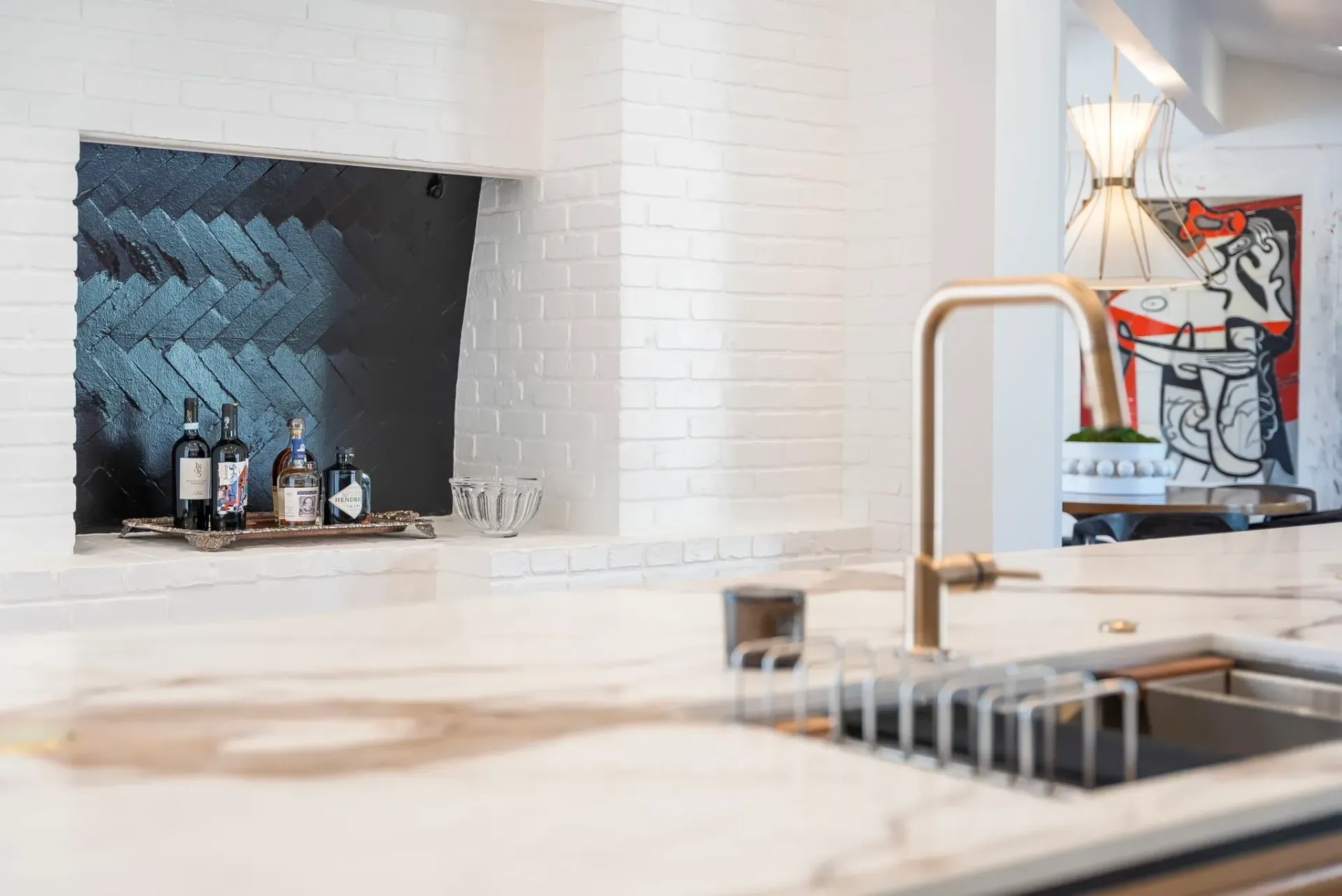Modern kitchen with white tile, marble island, brass faucet, wine bottles, and a small lamp display.