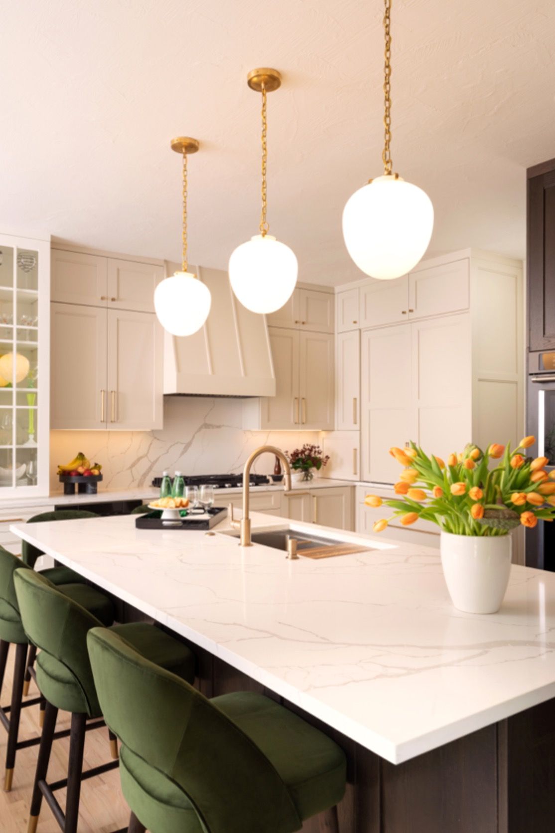 Modern white kitchen with marble island, green stools, brass pendant lights, and a vase of orange tulips