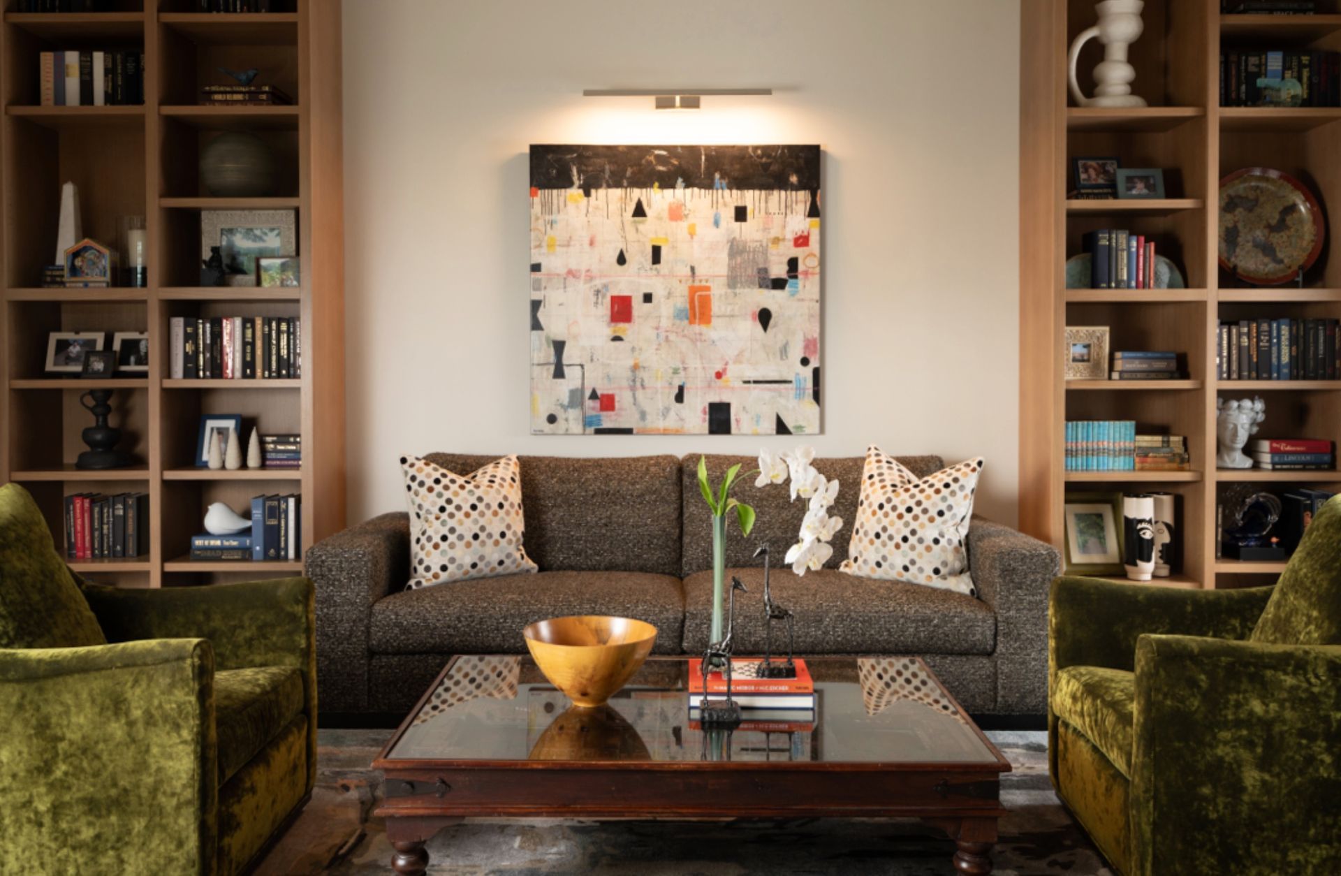 Cozy living room with gray sofa, green armchairs, bookshelves, coffee table, and abstract wall art