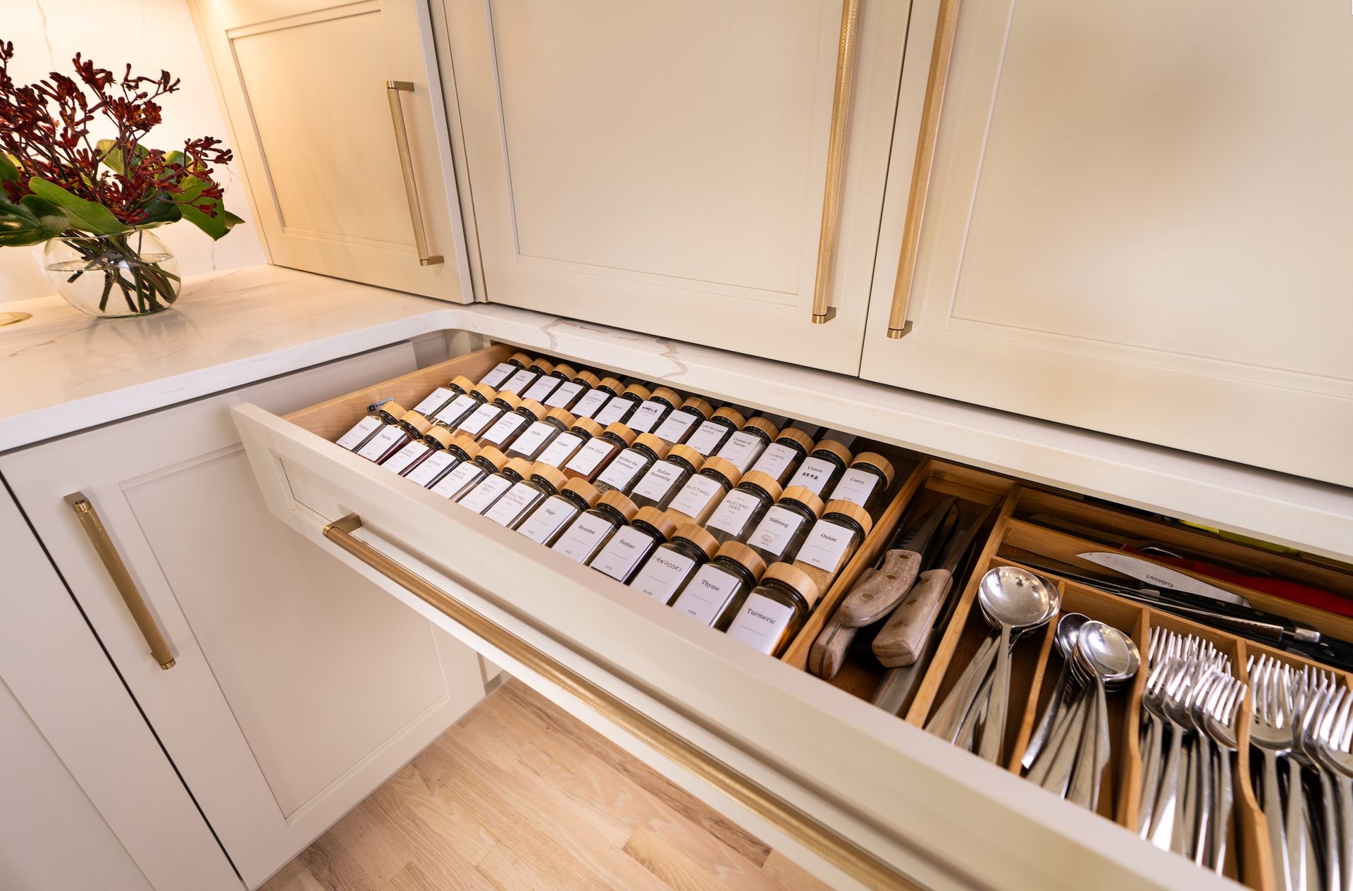 Open kitchen drawer with neatly organized spice jars and silverware next to a countertop with flowers