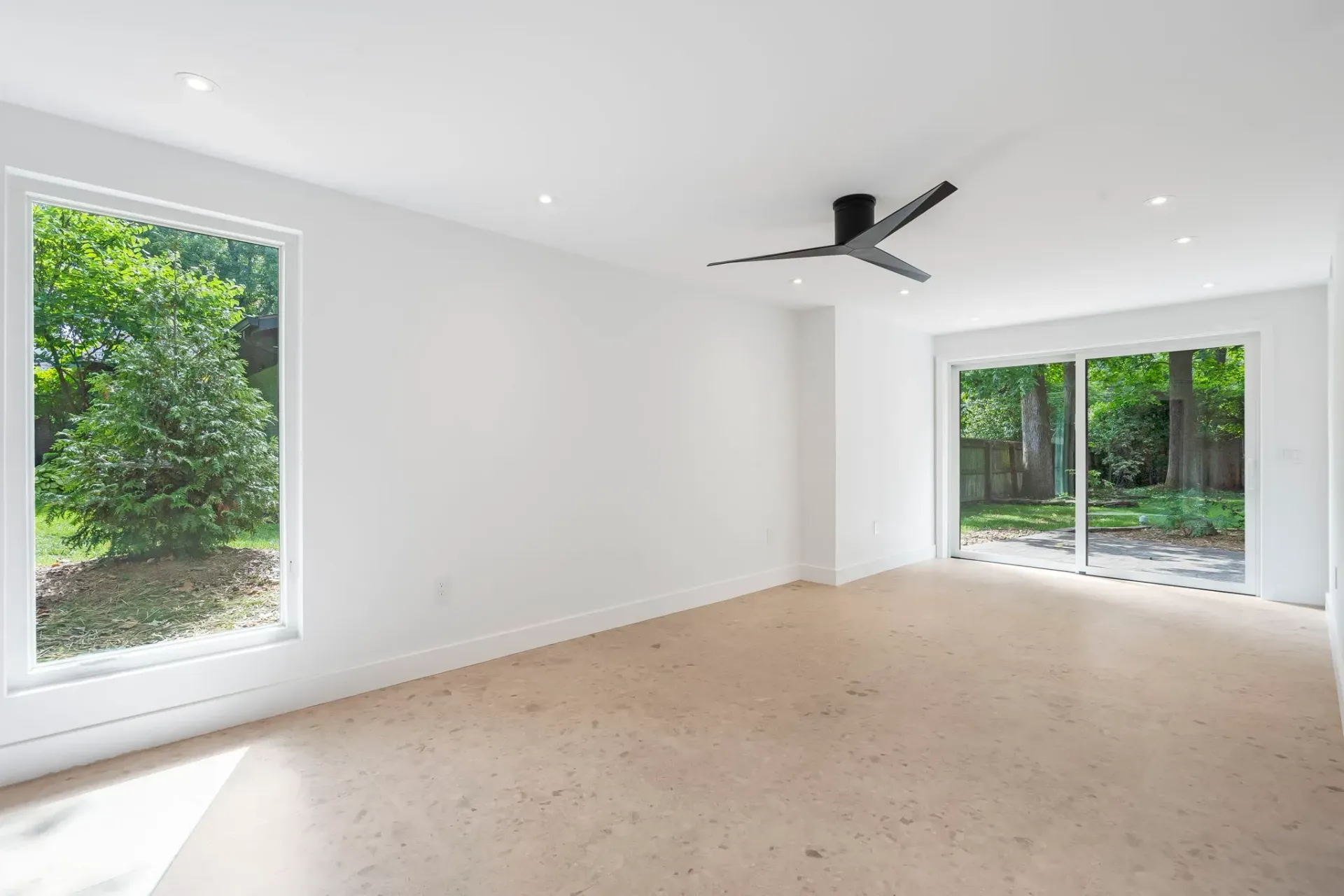 Bright empty living room with white walls, beige carpet, ceiling fan, and large windows overlooking greenery
