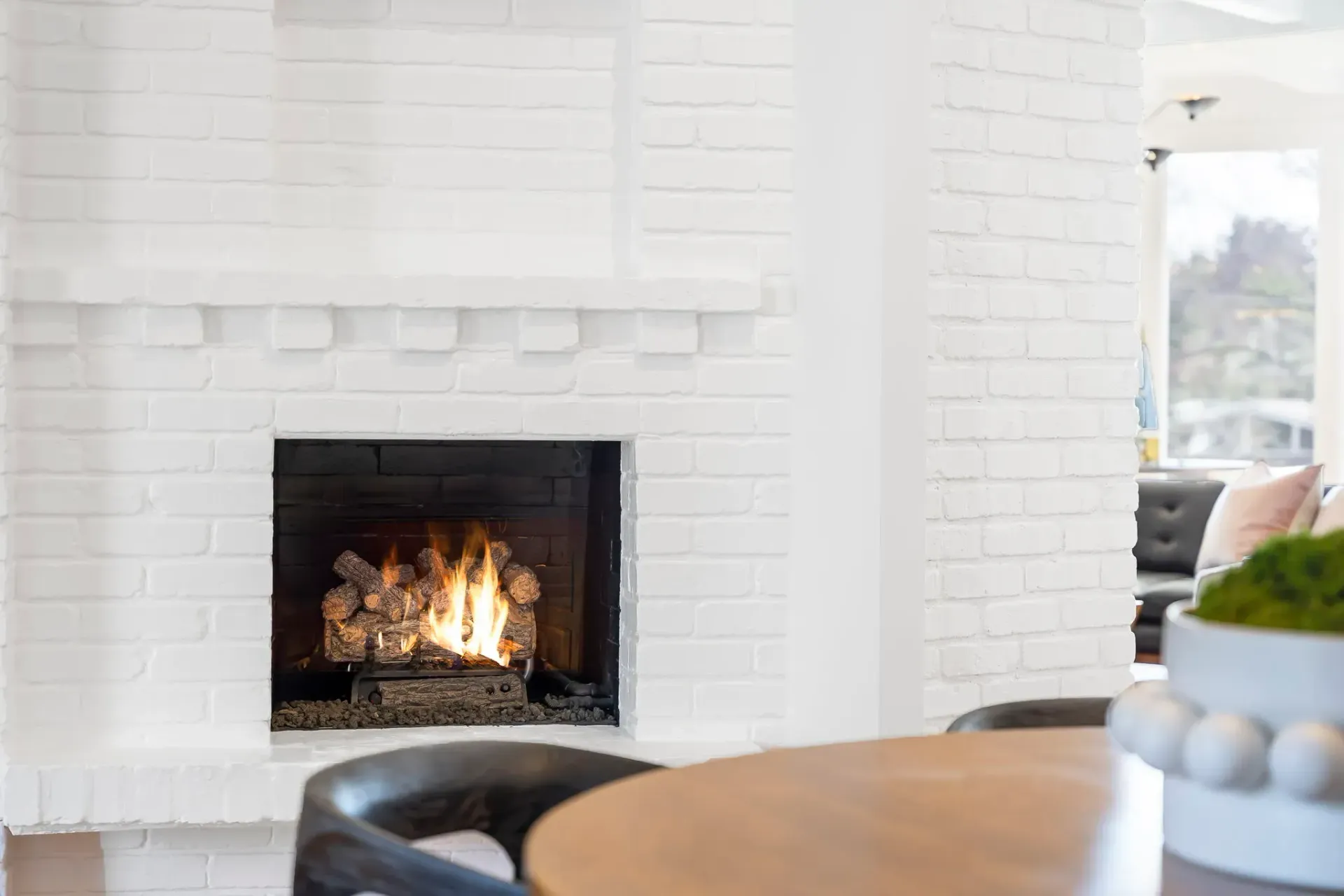 White brick fireplace with a lit fire in a bright living room beside a round table and black ottoman
