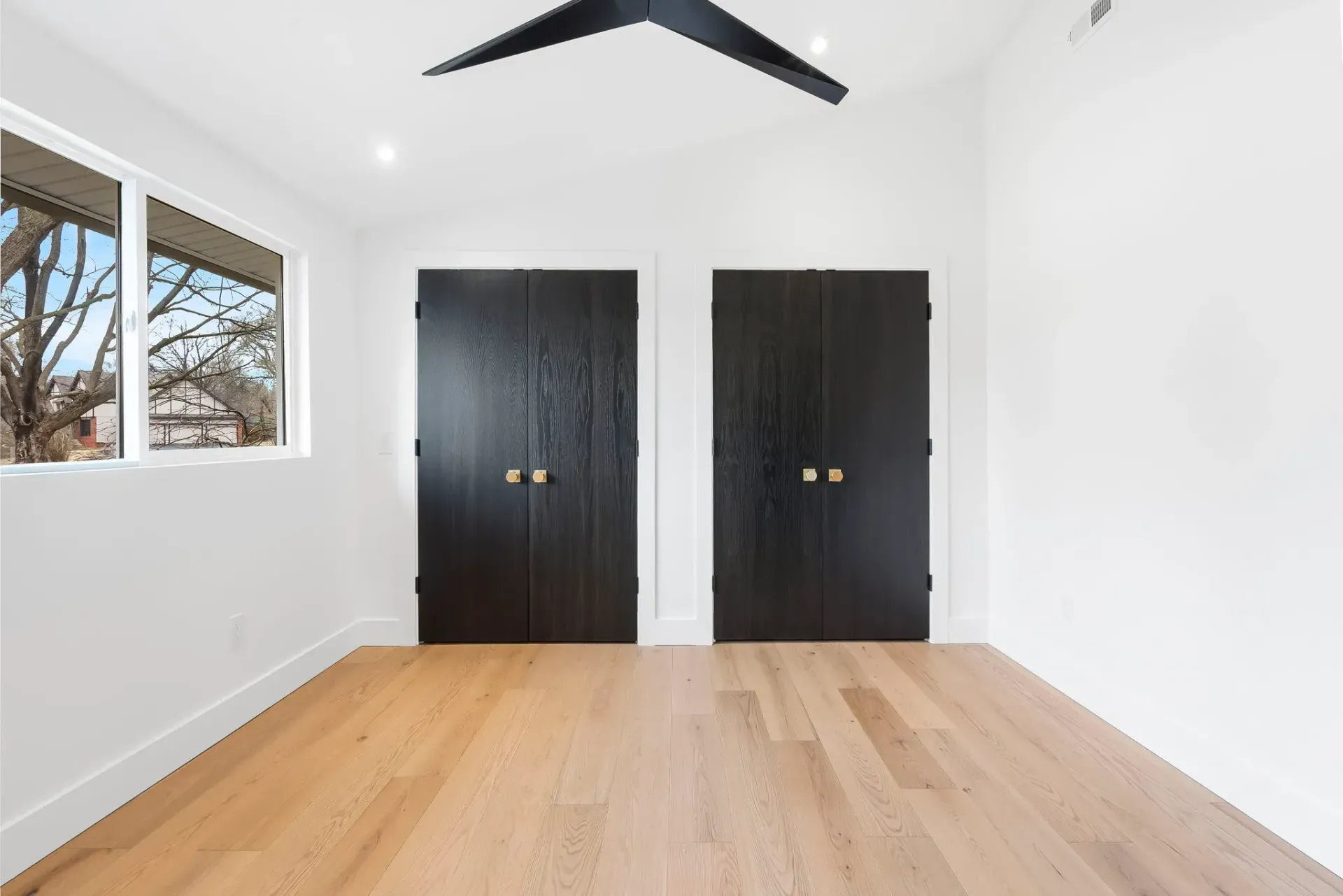 Empty modern bedroom with white walls, hardwood floor, black closet doors, and a window on the left