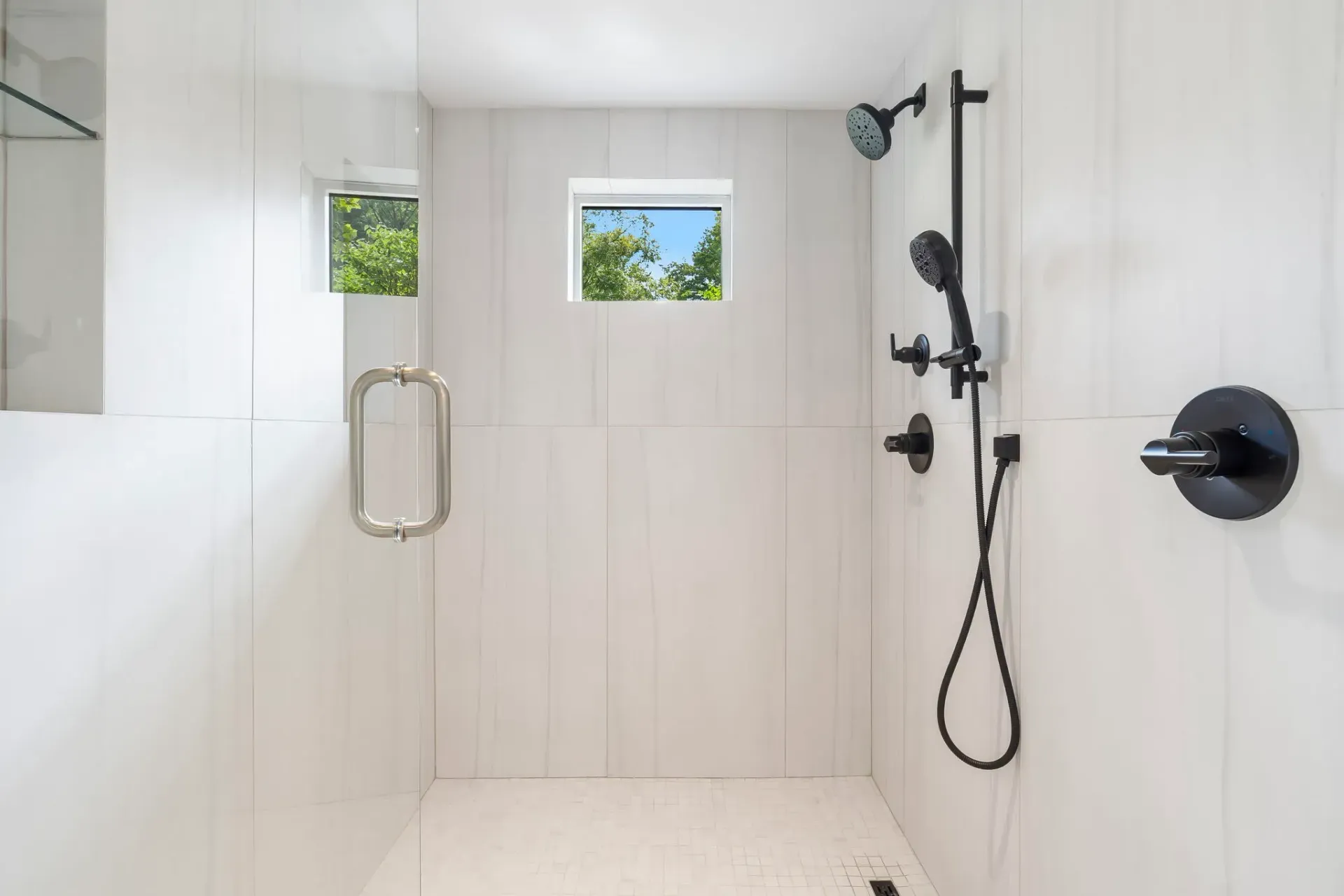 White tiled walk-in shower with black fixtures, a handheld showerhead, and small windows.