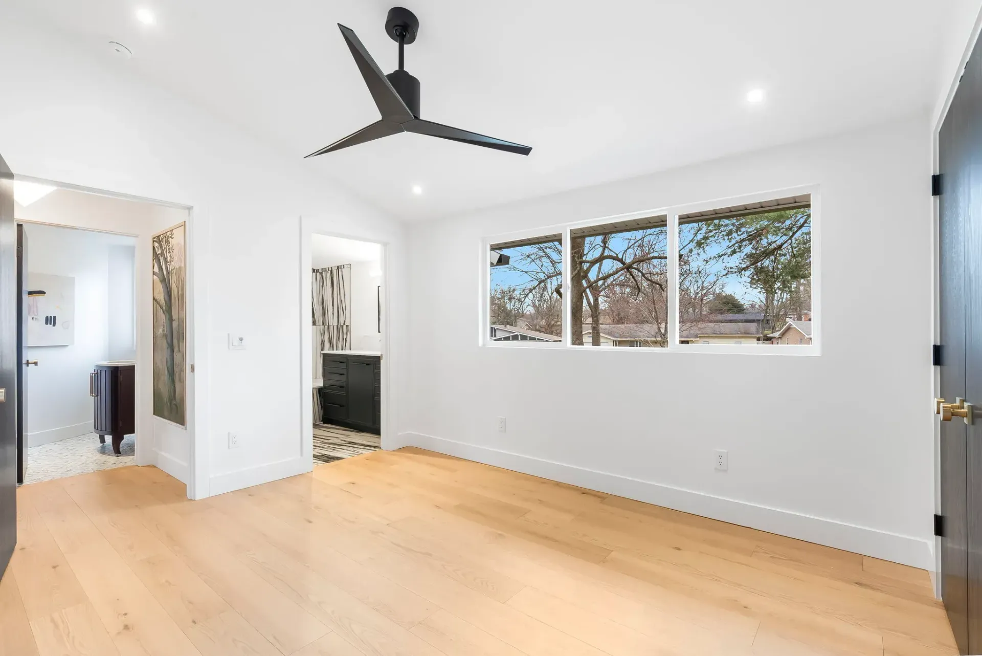 Bright empty bedroom with light wood floors, white walls, black ceiling fan, and windows overlooking trees