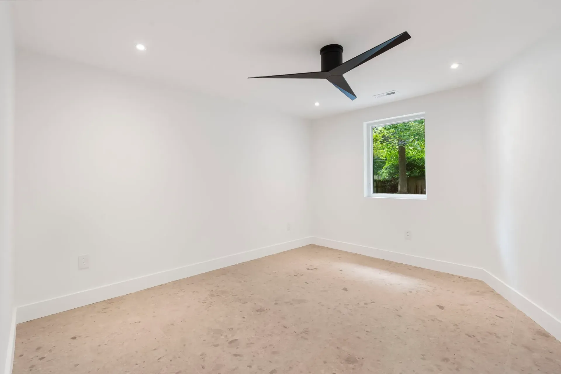 Empty white bedroom with beige carpet, black ceiling fan, and a small window showing greenery