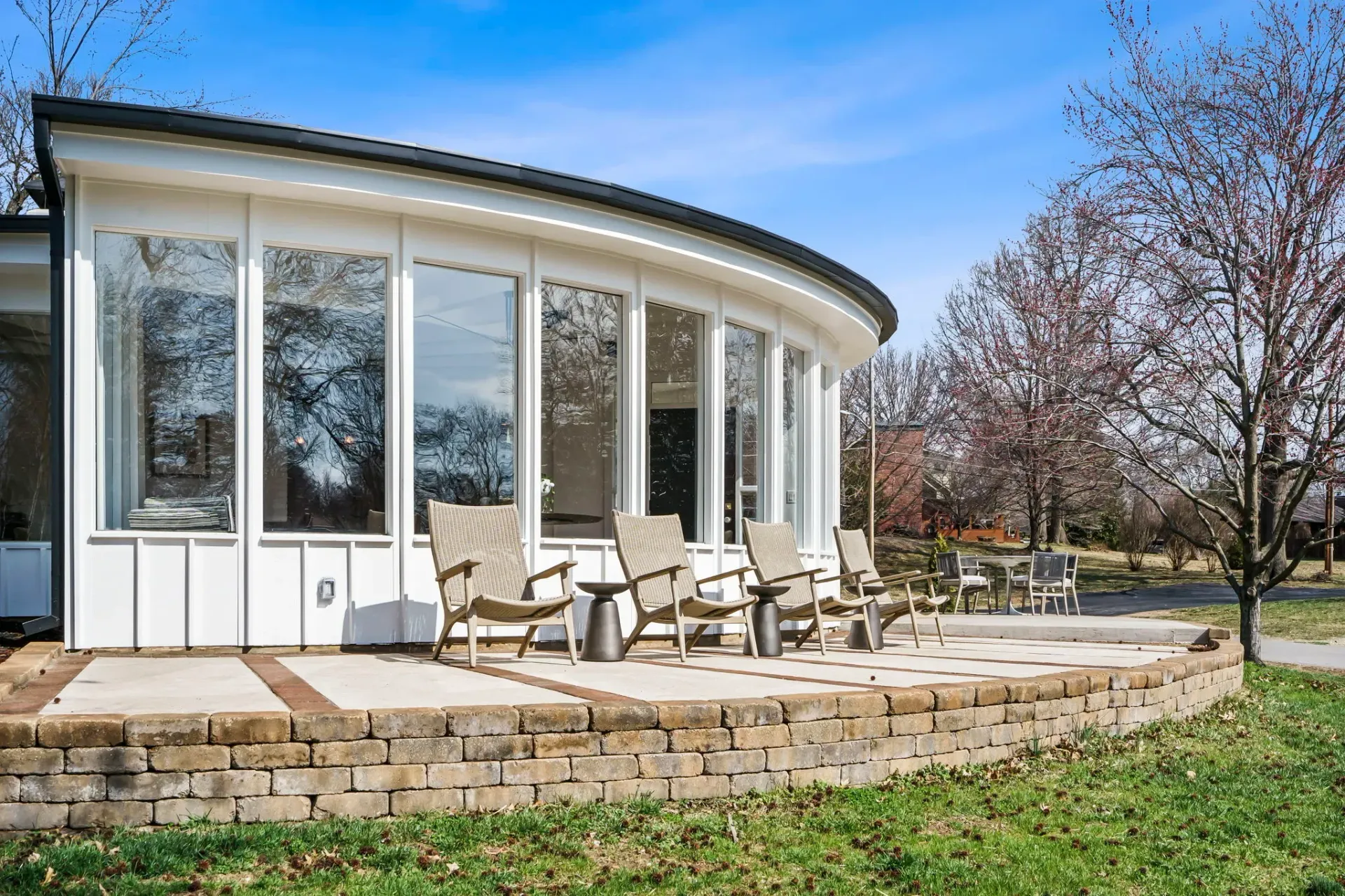 Curved glass building with patio chairs on a stone terrace beside a grassy lawn and trees