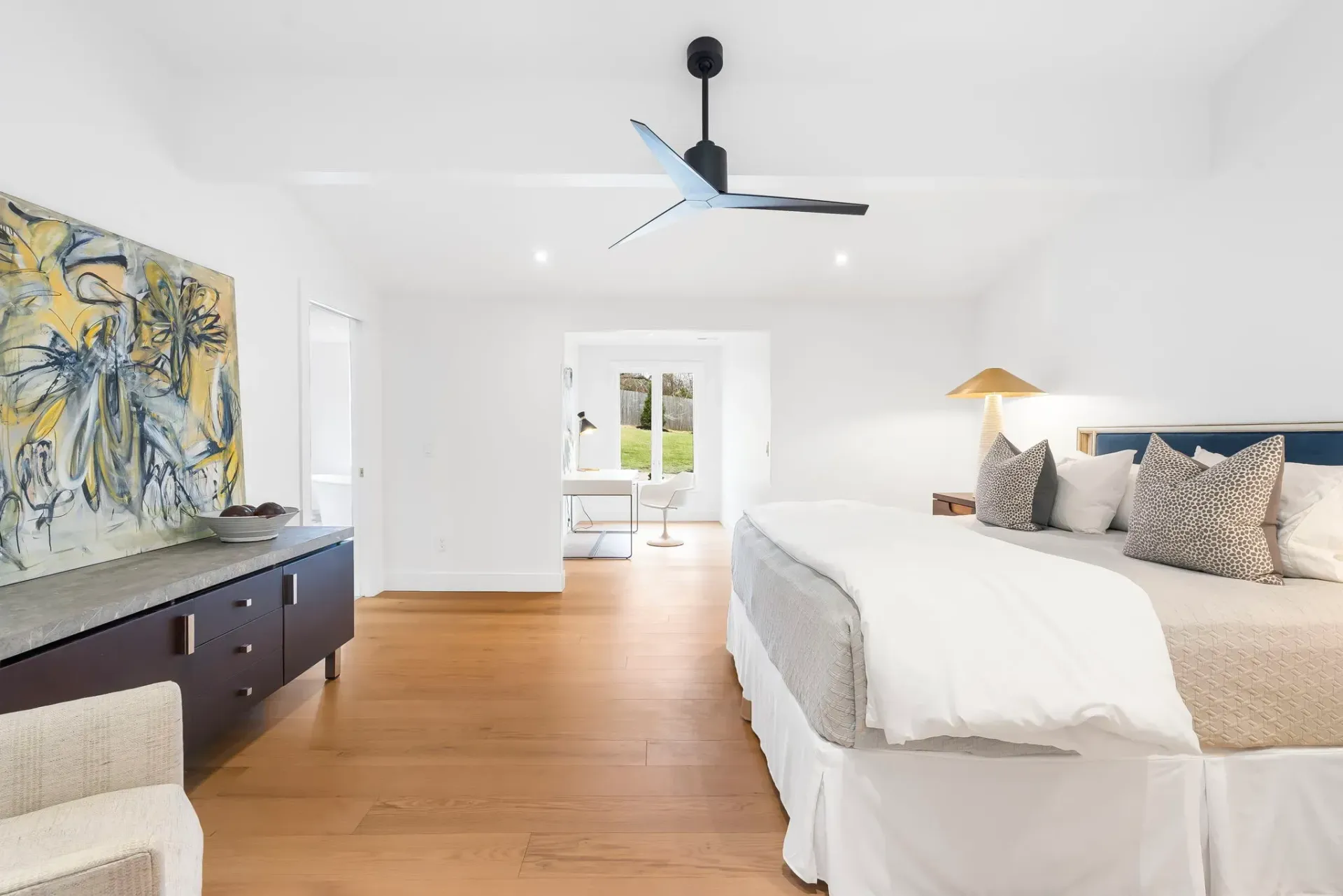 Bright minimalist bedroom with white bedding, wood floors, abstract art, and a ceiling fan