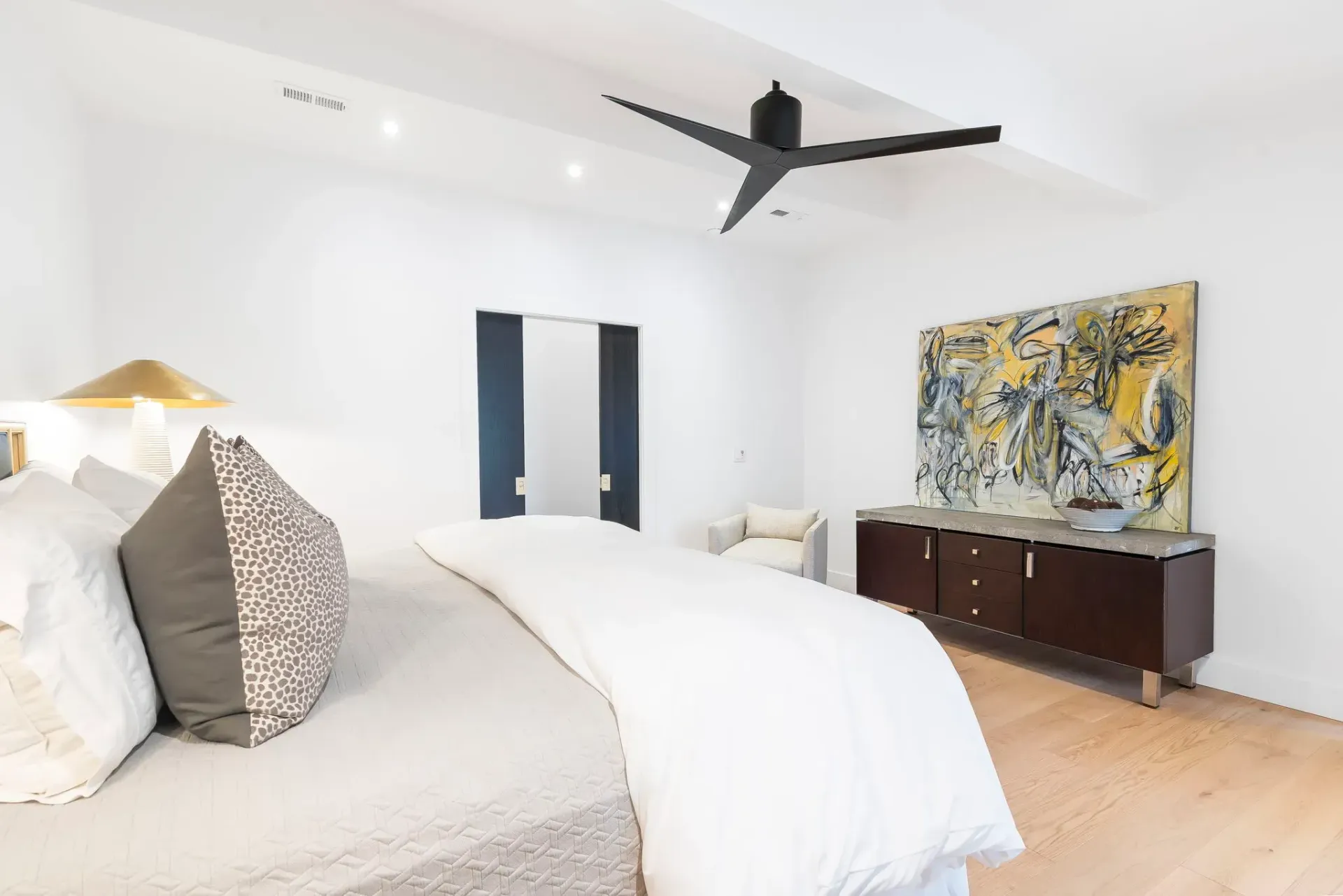 Bright bedroom with a bed, ceiling fan, and colorful abstract art above a dark dresser.