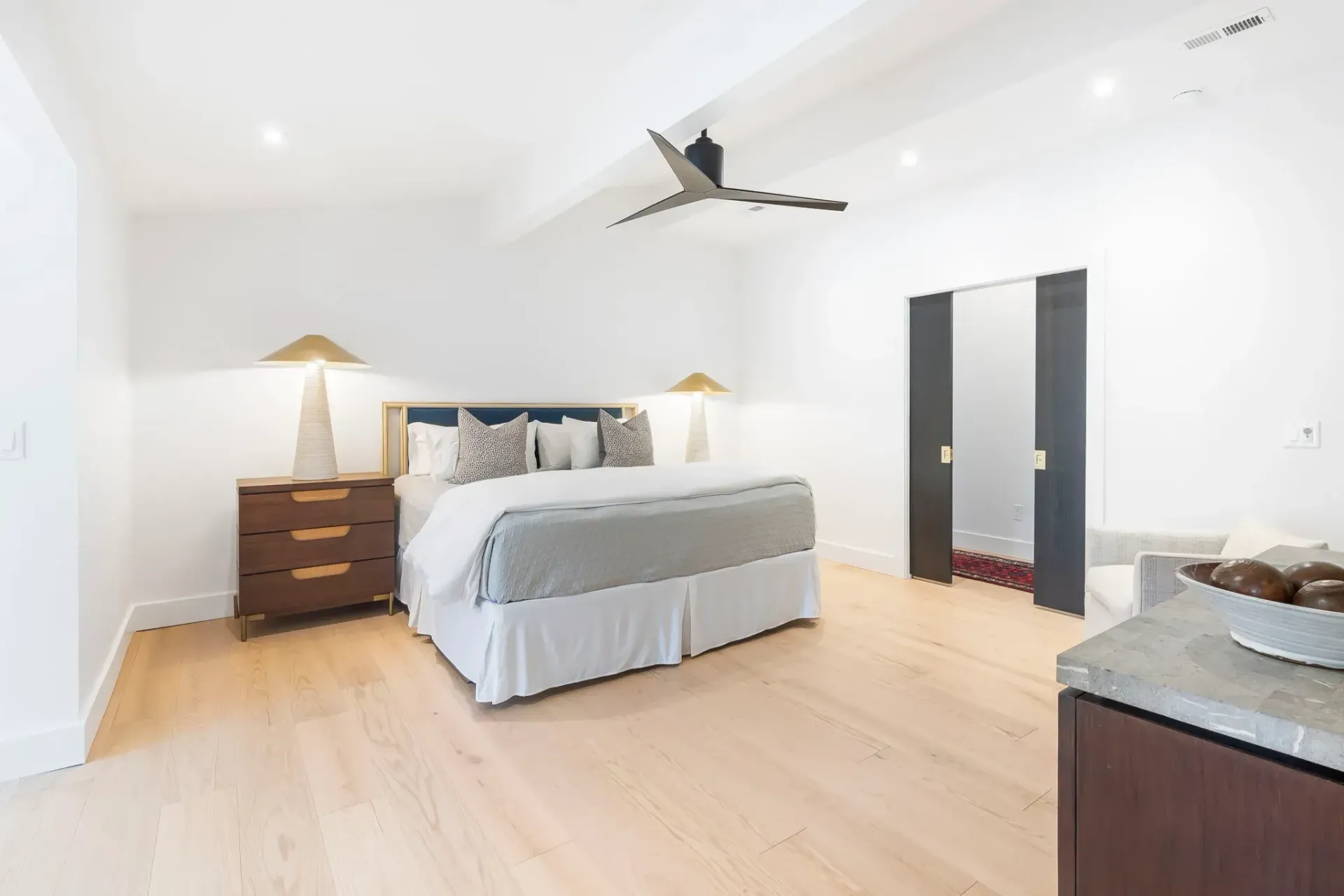 Bright bedroom with white walls, wood floor, gray bed, bedside lamps, and dark closet doors