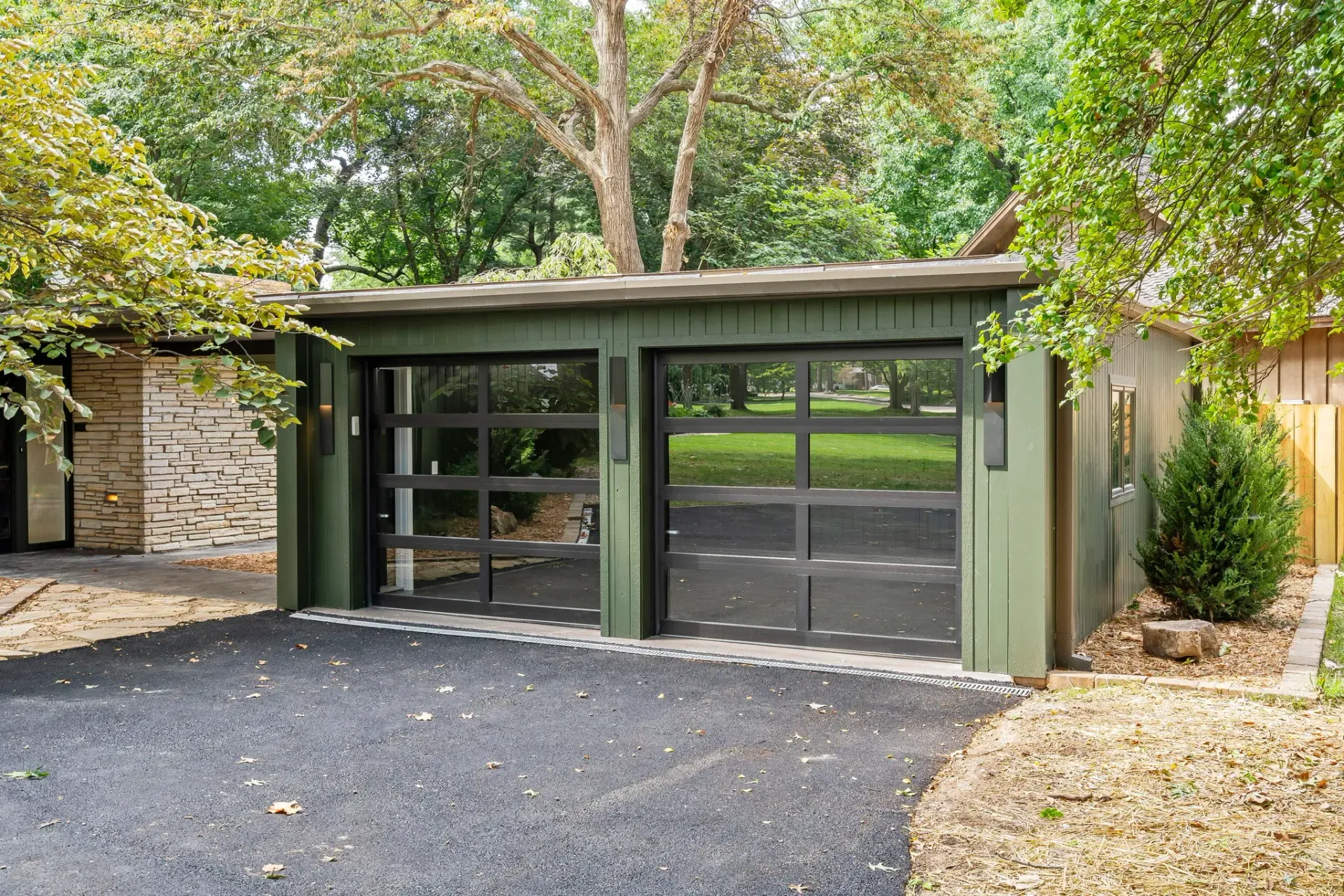 Modern green garage with glass doors beneath trees, gravel driveway in front