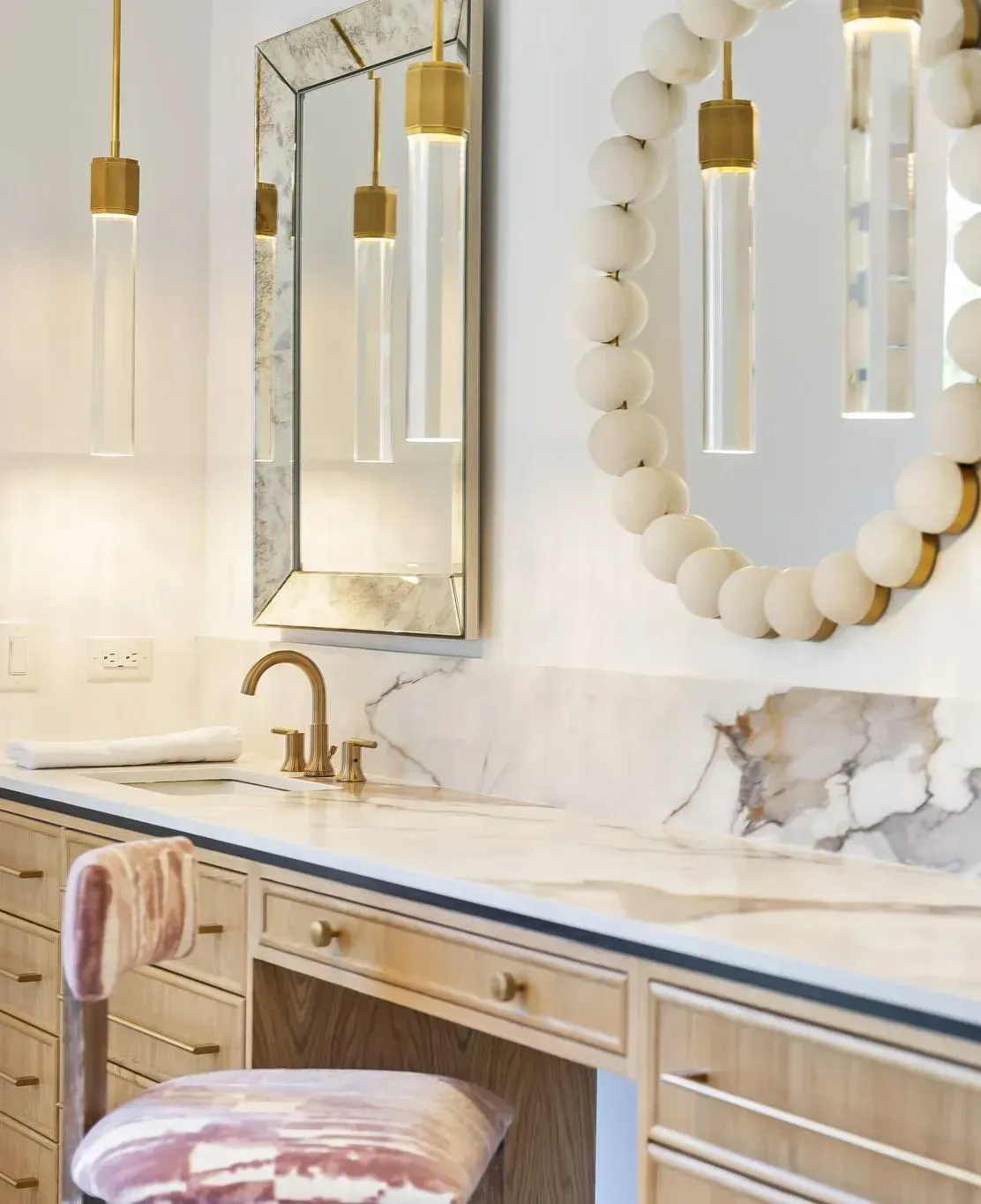 Elegant bathroom vanity with gold fixtures, marble countertop, round mirror, and pink stool under pendant lights