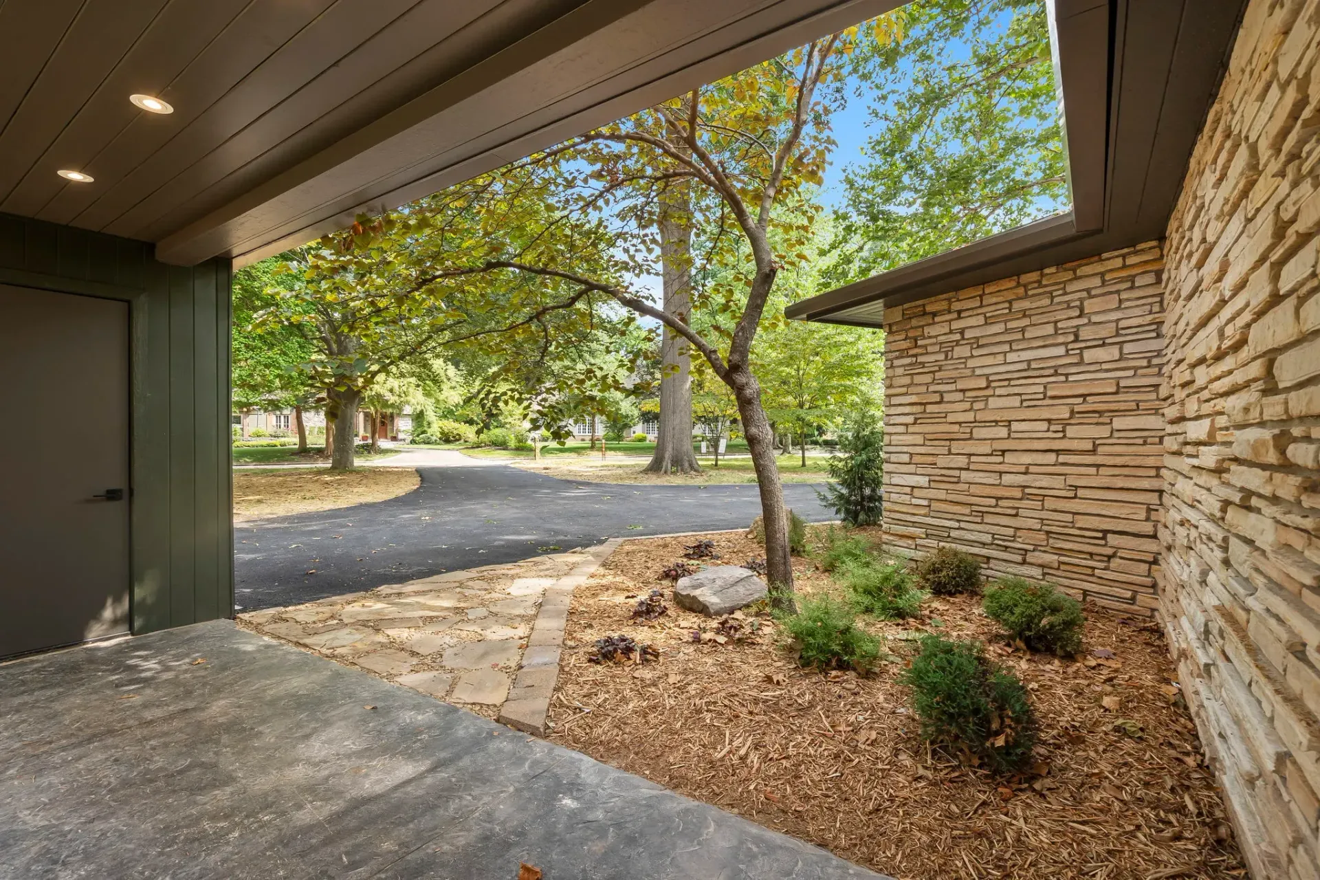 Covered patio with stone wall, gravel yard, and sunlit trees beyond