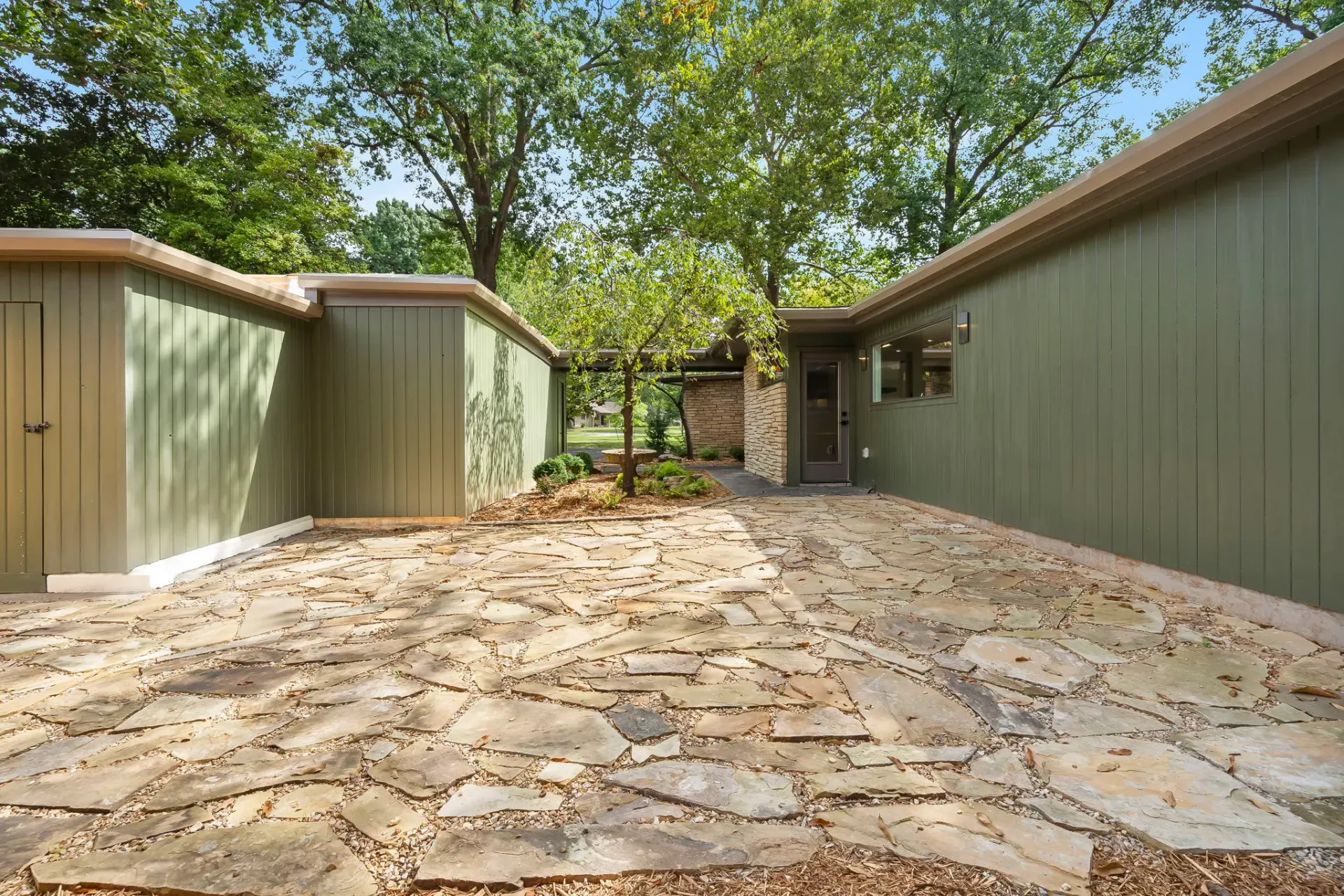Stone patio between green buildings with trees overhead and a doorway at the far end