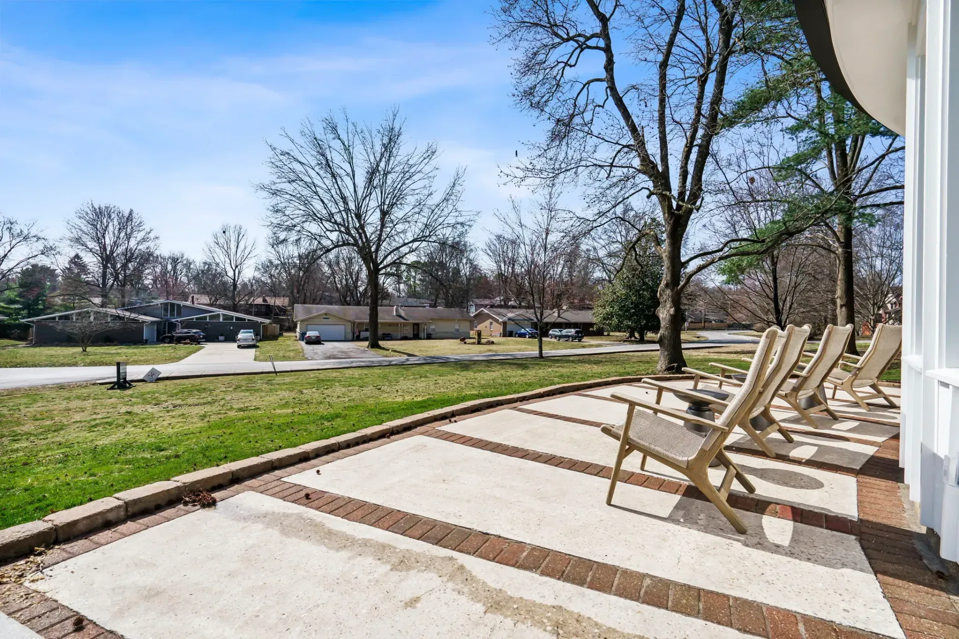 Row of tan lounge chairs on a sunny patio beside a grassy park and trees
