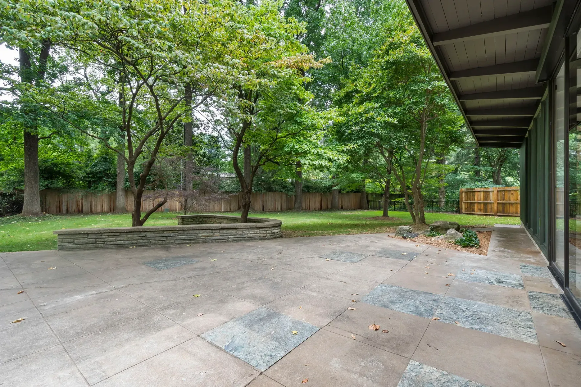 Empty outdoor patio beside a green tree-lined yard, with a covered roof on the right and gravel patches on the ground