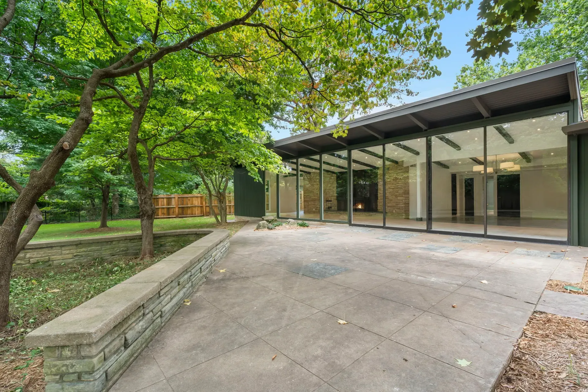 Modern house with glass walls and a covered patio beside a tree-lined gravel driveway
