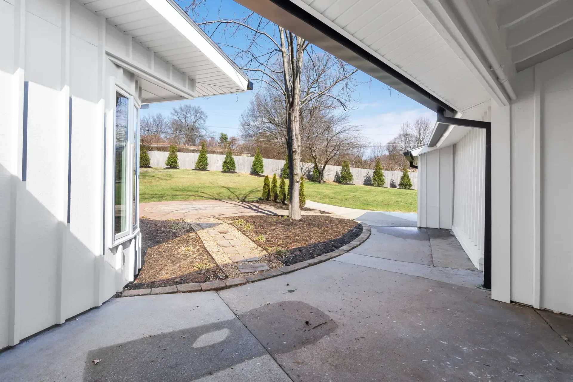 Covered concrete walkway leading to a yard with trees and landscaping.