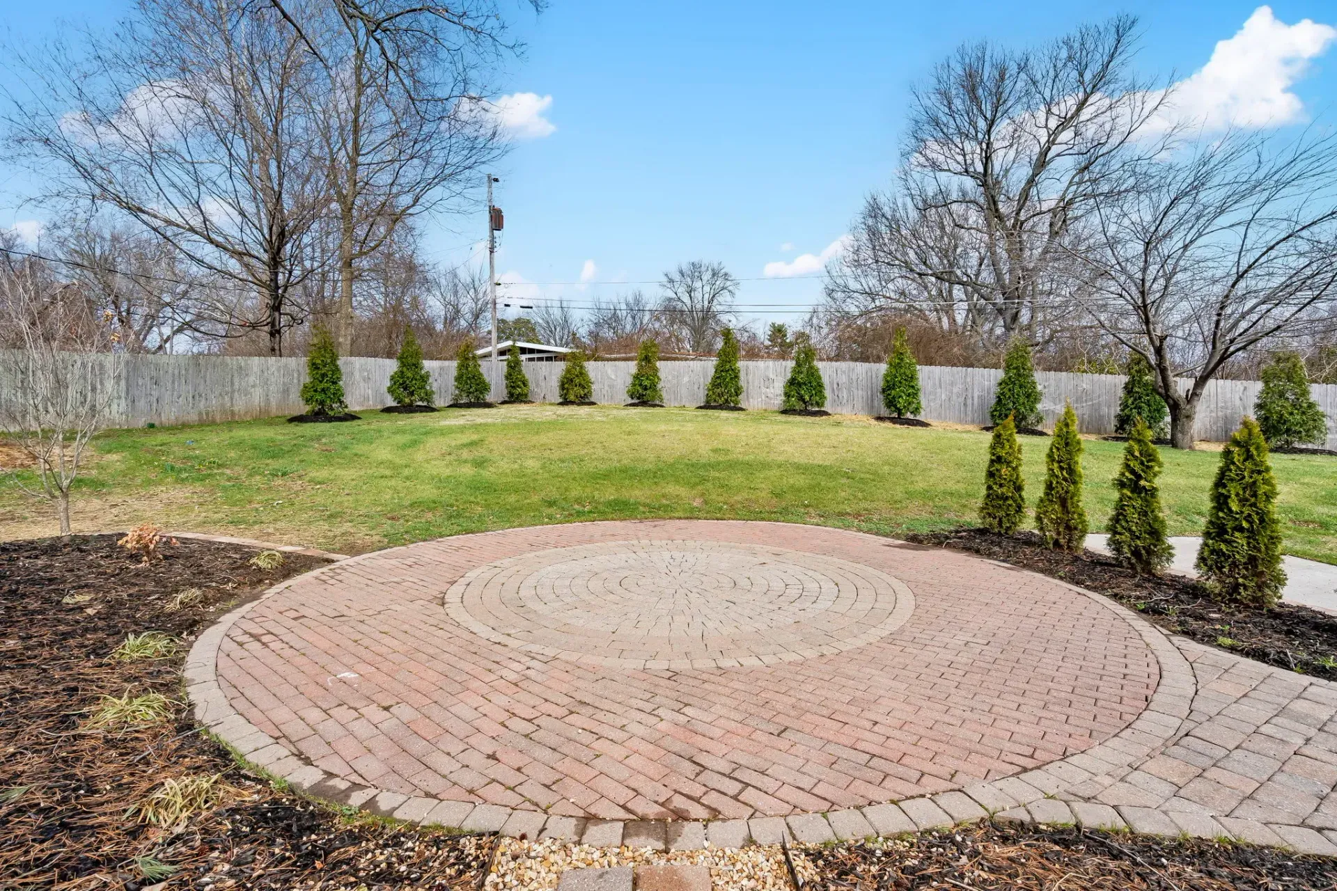 Circular brick patio in a landscaped backyard with grass, shrubs, and leafless trees under a blue sky