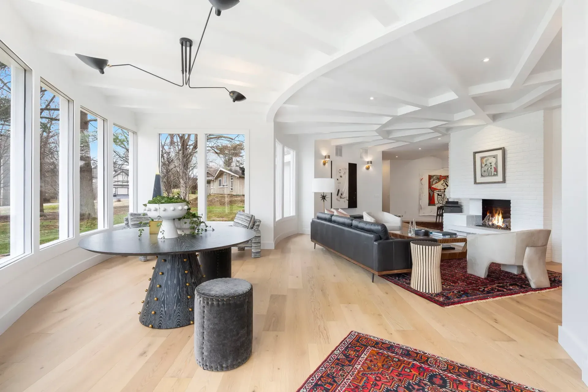 Bright modern living-dining room with large windows, black table and sofas, light wood floors, and red rugs