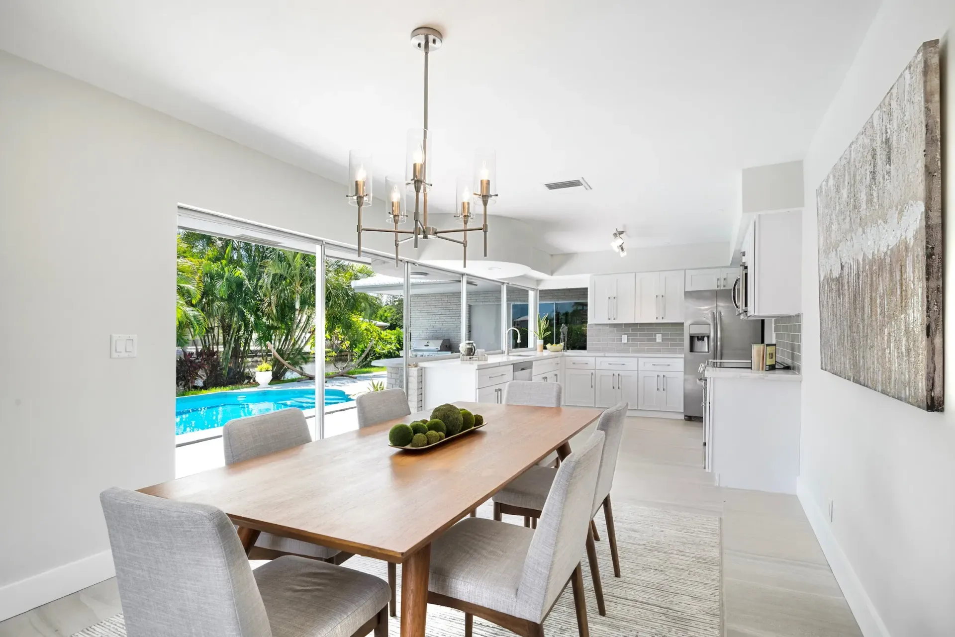 Bright dining room with wooden table, gray chairs, chandelier, and sliding doors opening to a pool and greenery