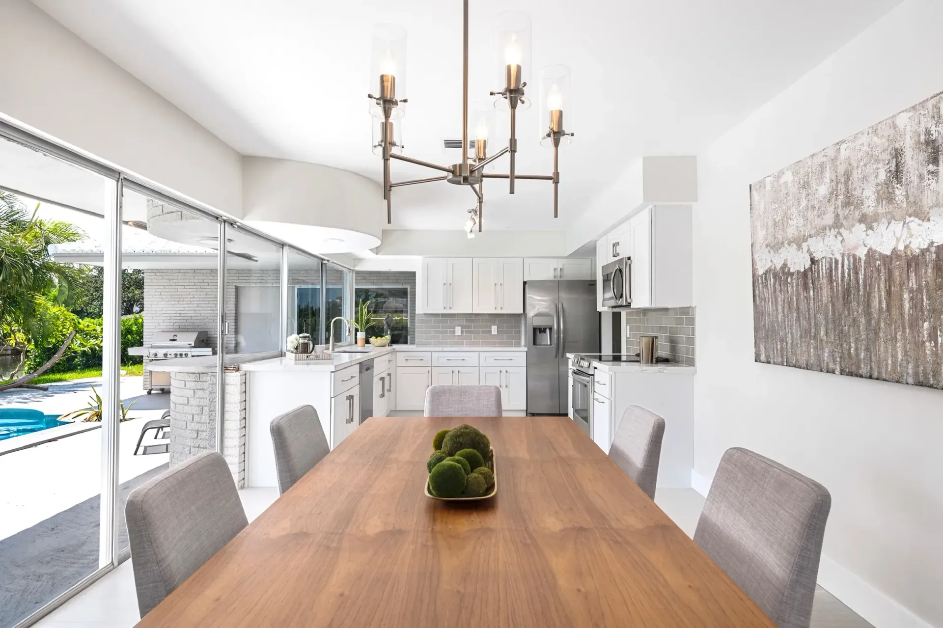 Bright modern dining room with wooden table, gray chairs, chandelier, and open view to kitchen and pool.