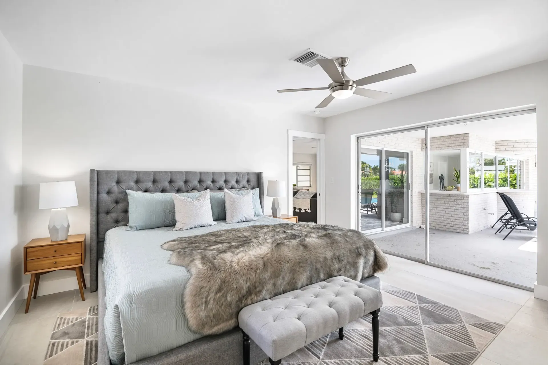 Bright bedroom with tufted bed, bench, and sliding doors to a sunroom with patio chairs