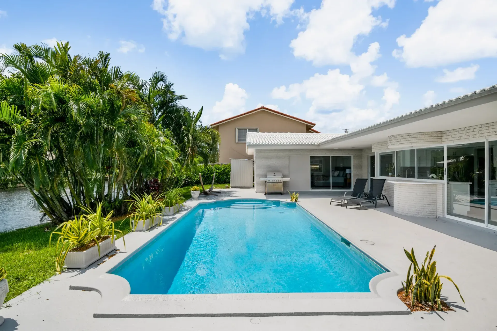 Modern backyard with a turquoise swimming pool, lounge chairs, and a white house under a blue sky.