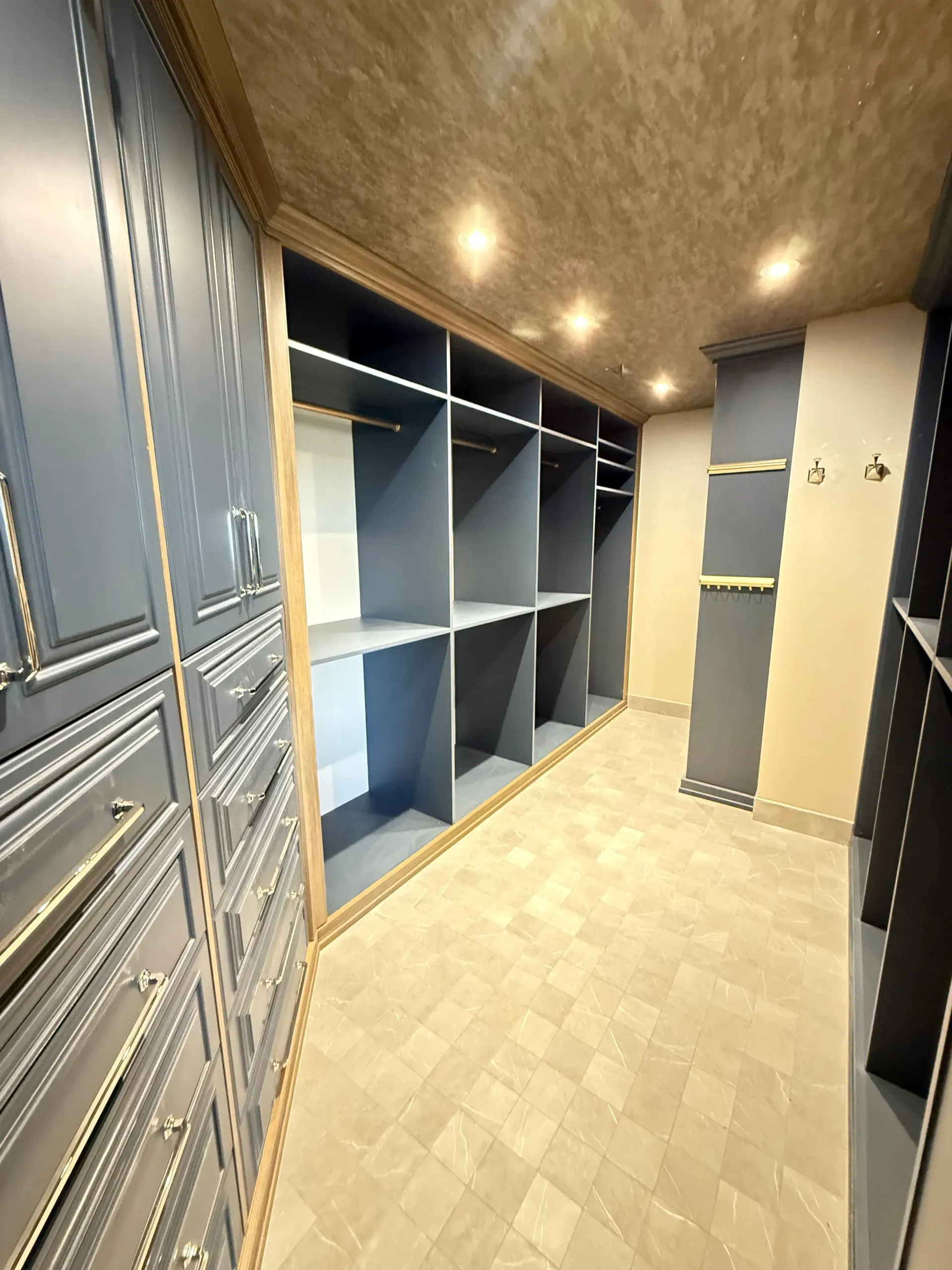 Empty walk-in closet with blue shelves, gray cabinets, and warm ceiling lights.