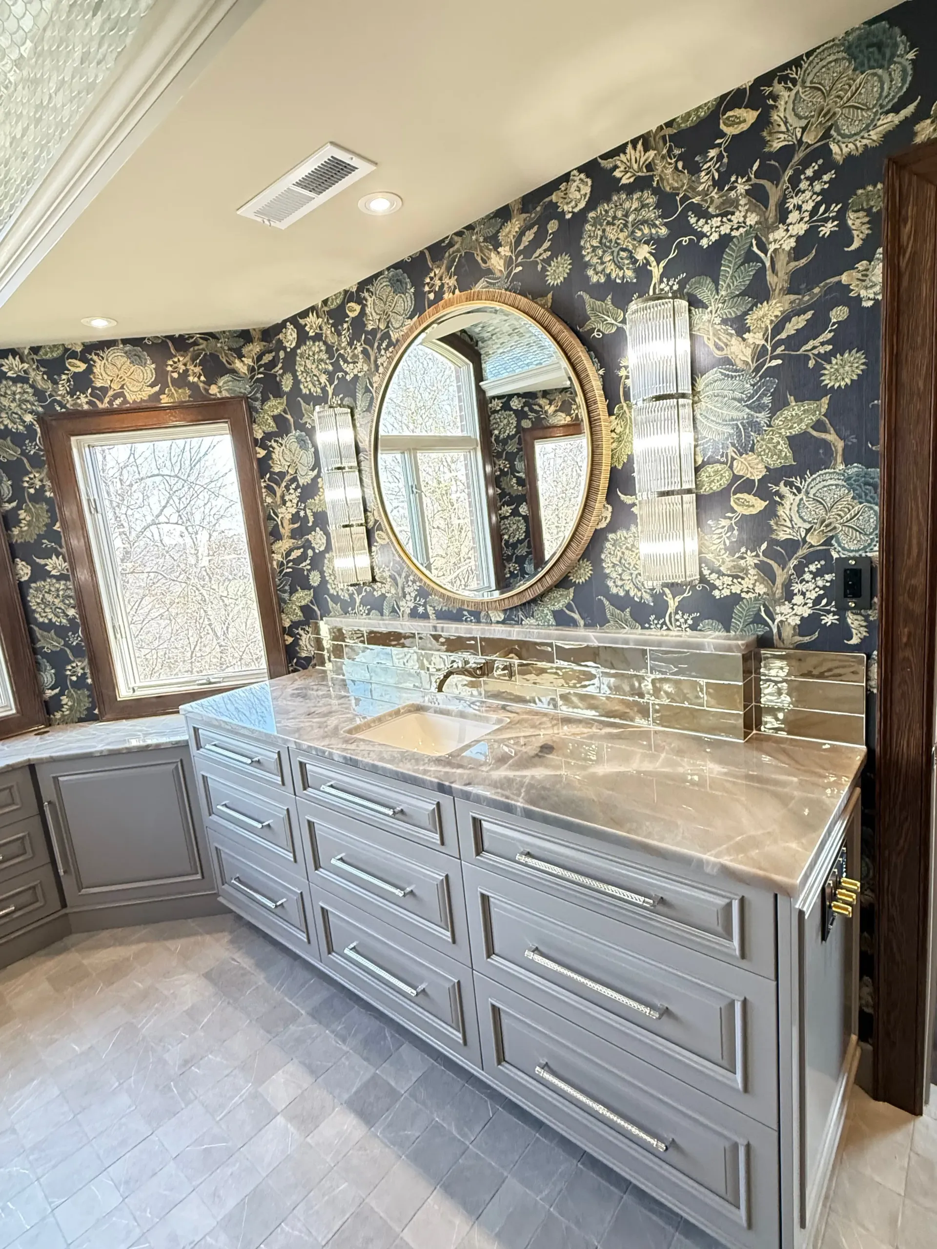 Bathroom vanity with gray cabinets, marble countertop, round mirror, and floral black-and-gold wallpaper