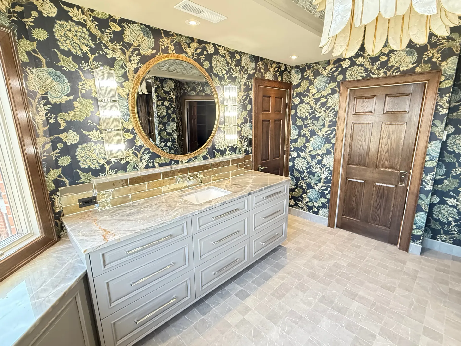 Bathroom vanity with gold floral wallpaper, round mirror, double sink, and wooden doors