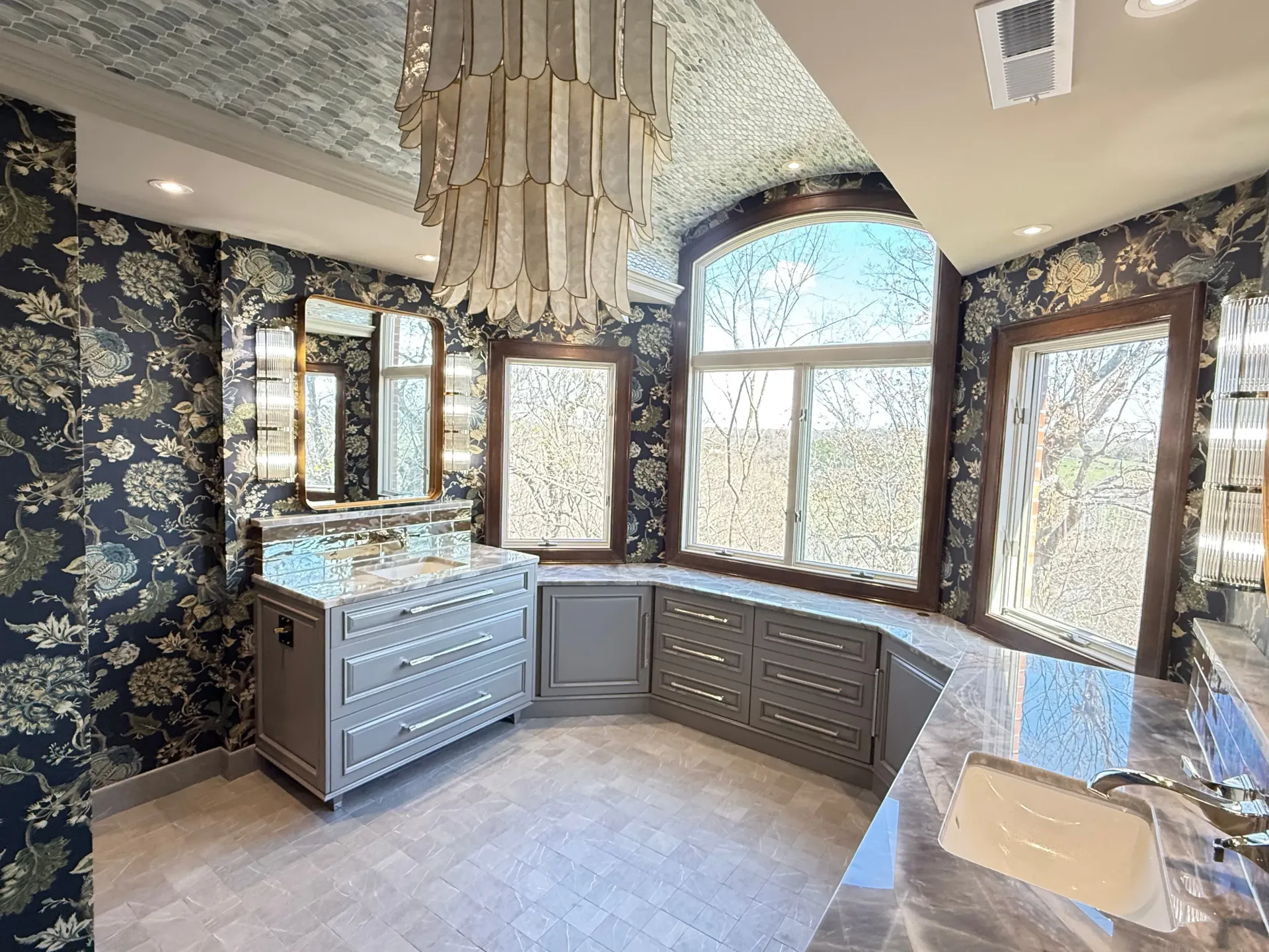 Elegant bathroom with floral wallpaper, chandelier, marble vanity, and large arched windows.