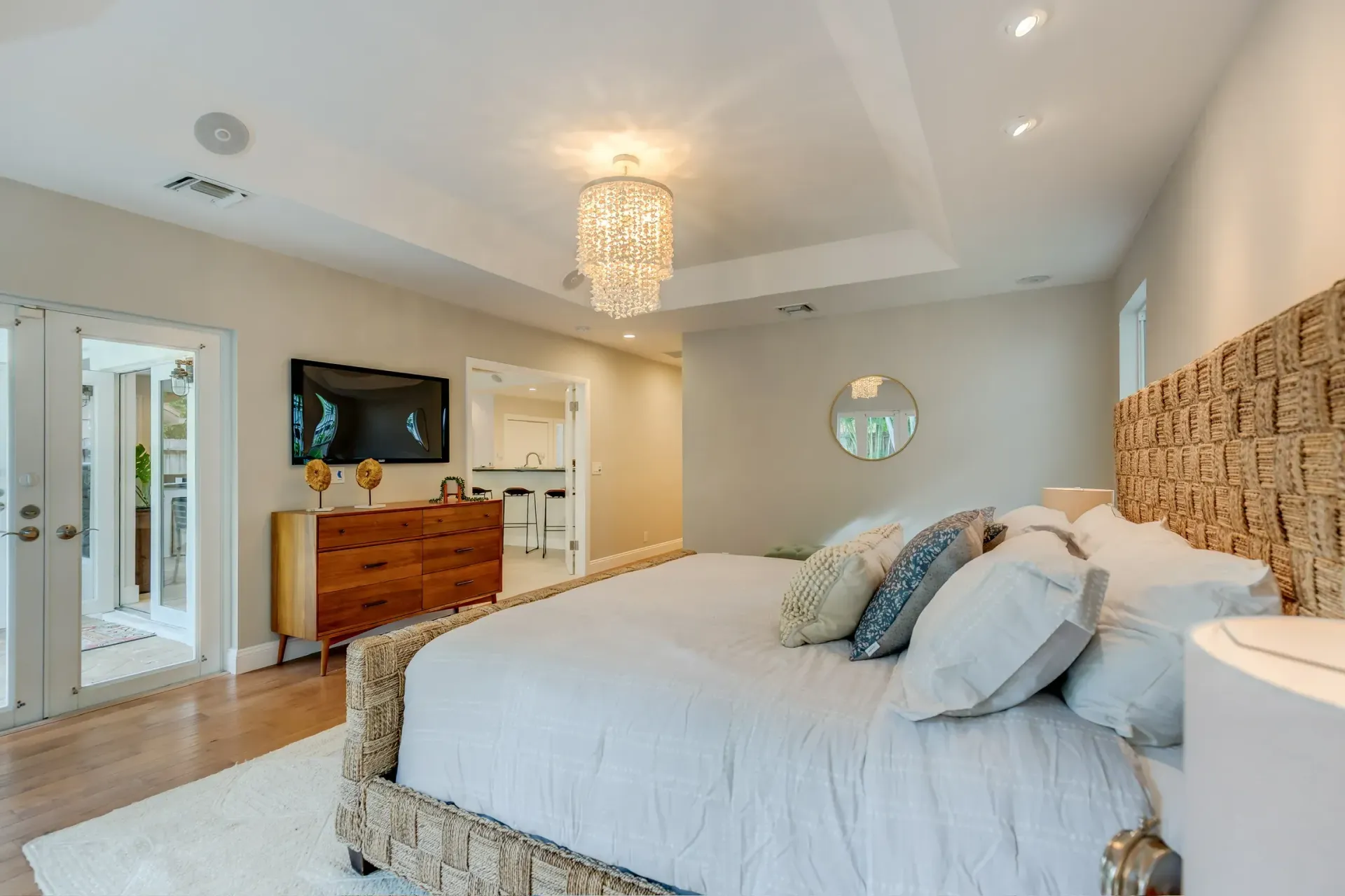Bright bedroom with a large bed, chandelier, dresser, wall-mounted TV, and mirrored closet doors