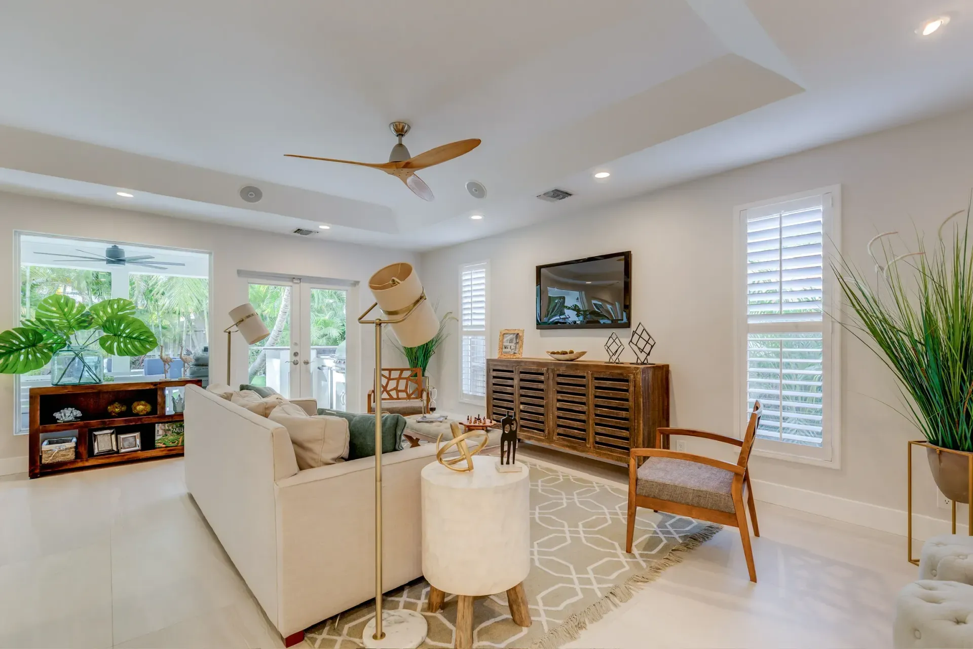 Bright living room with white sofas, wooden TV console, ceiling fan, and large windows