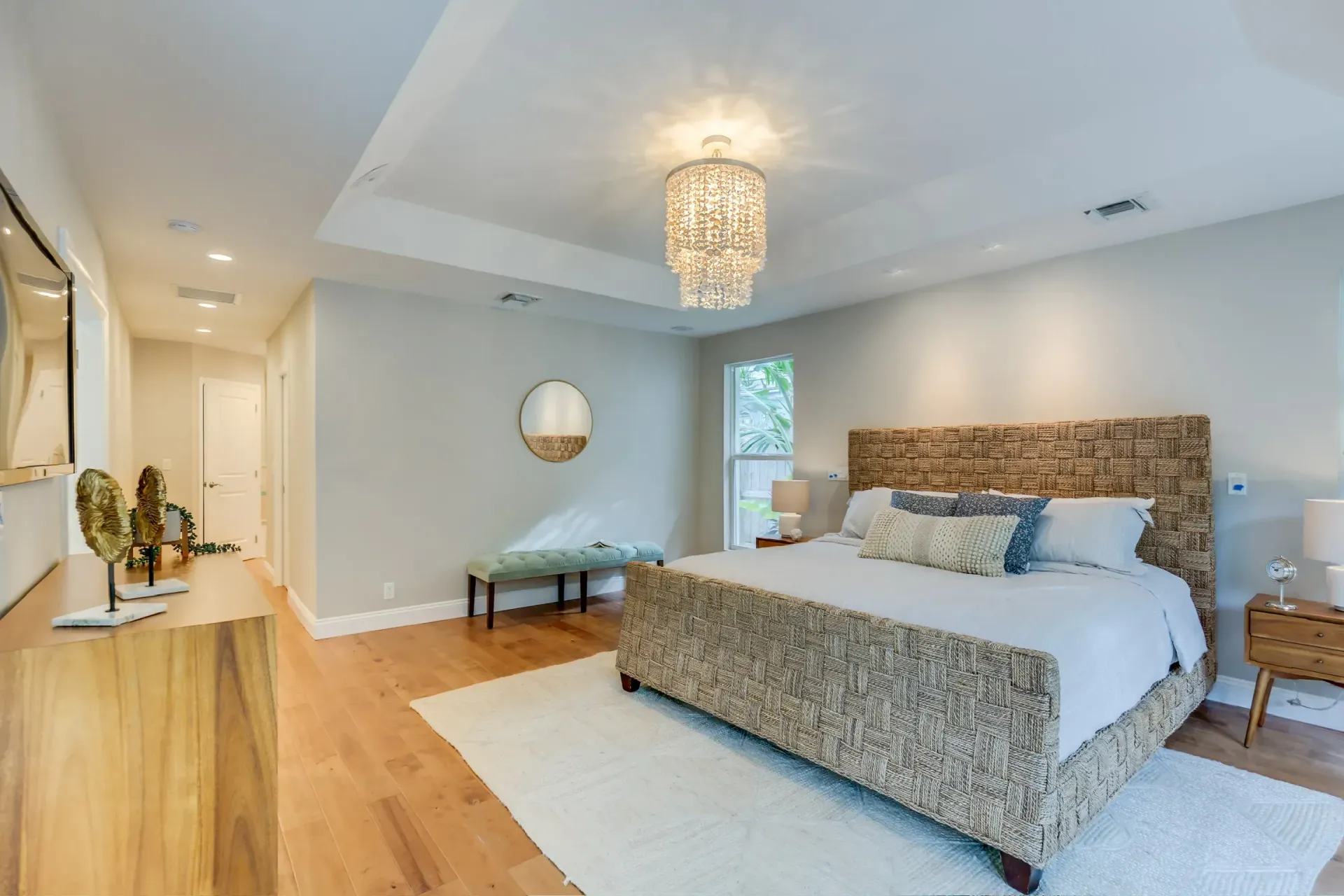 Bright modern bedroom with a large bed, chandelier, hardwood floors, and a bench by the window.