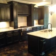 Dark kitchen with black cabinets, marble island, stainless stove, and warm pendant lighting