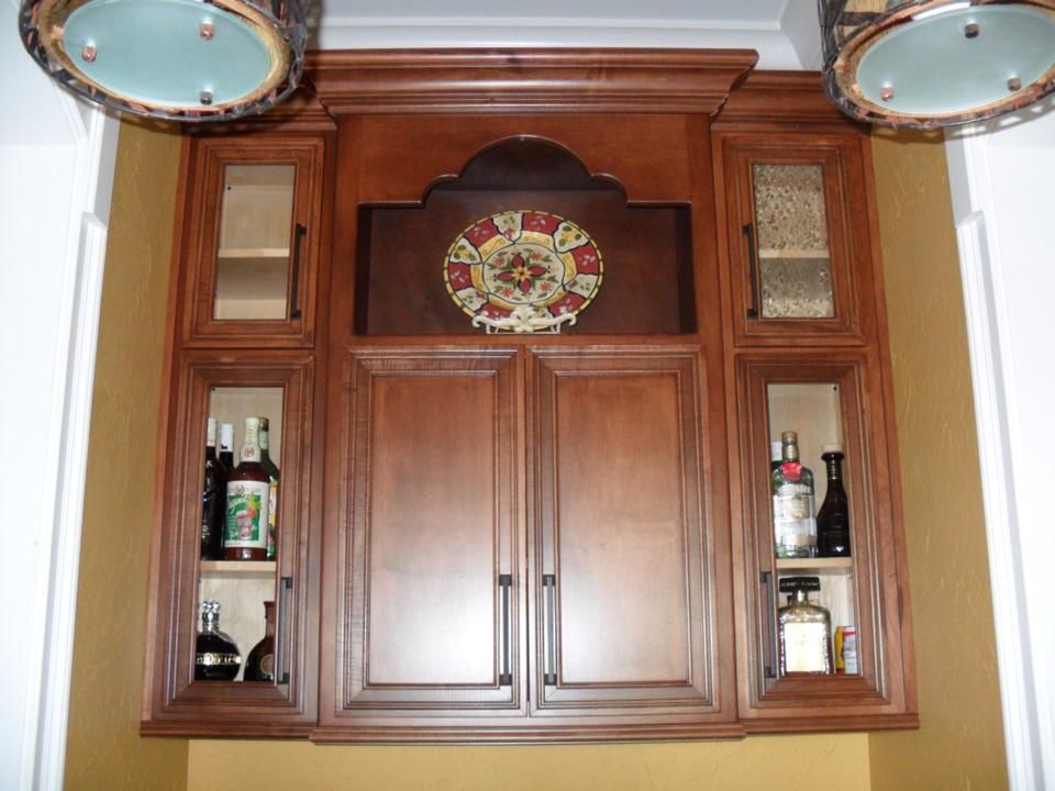 Wooden bar cabinet with glass shelves, bottles, and decorative crest on a white wall