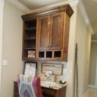 Wooden wall cabinet above a desk in a hallway, with open shelves and a chair beneath it.