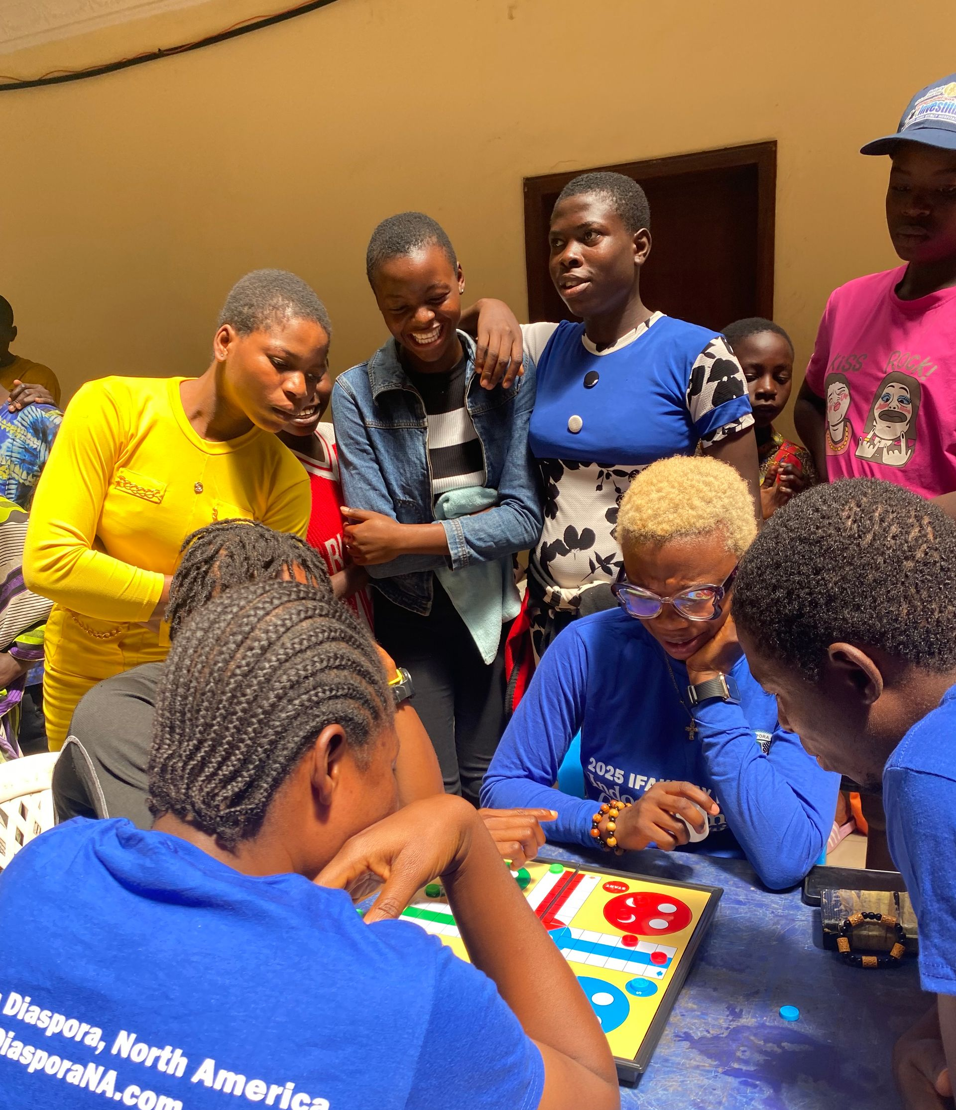 People playing a board game together at a table. Several people are watching and smiling.