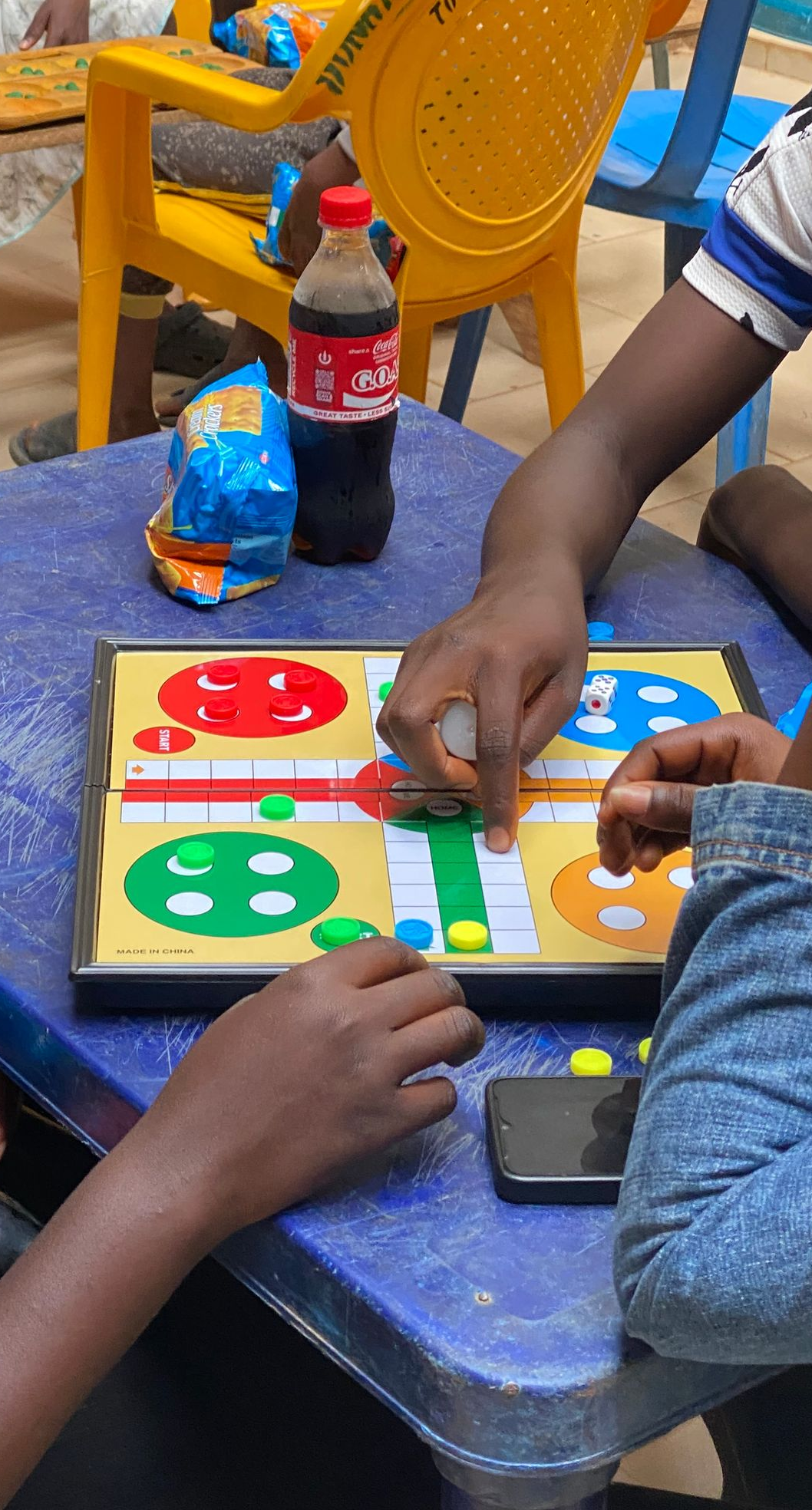 People playing Ludo board game at a table, with a soda bottle and snack bag nearby.