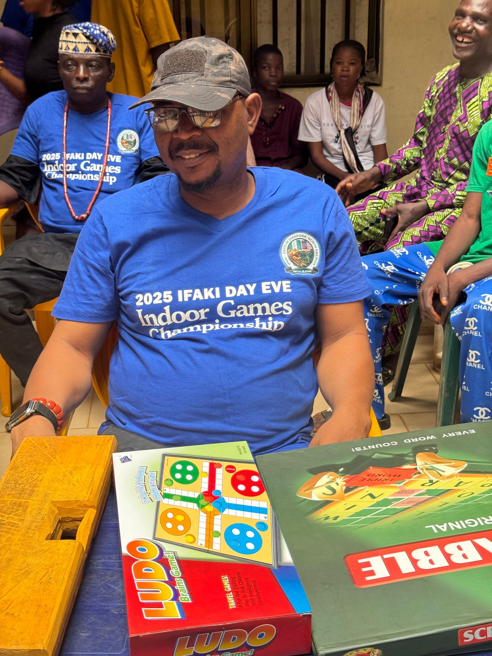 Man smiles, sits with board games: Ludo, Scrabble. Blue shirt: